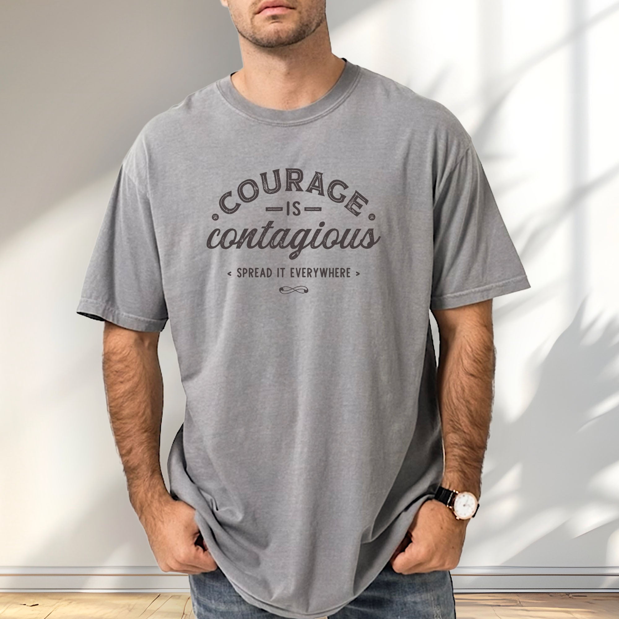 Male model wearing grey vintage style typography shirt reading "Courage Is Contagious Spread It Everywhere" in distressed lettering.