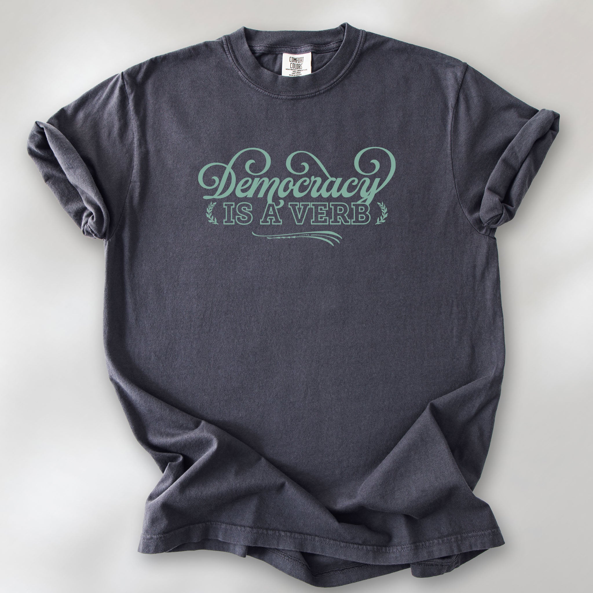 Graphite t-shirt with the phrase “Democracy Is a Verb” in decorative mint green lettering.