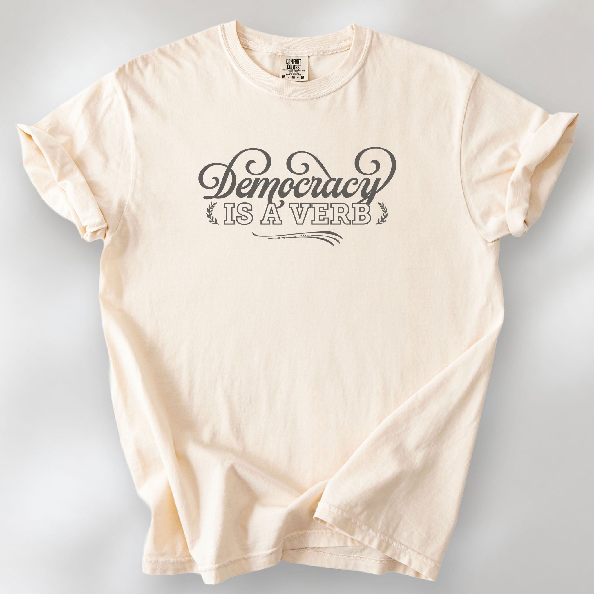 Ivory t-shirt with the phrase “Democracy Is a Verb” in decorative mint green lettering.