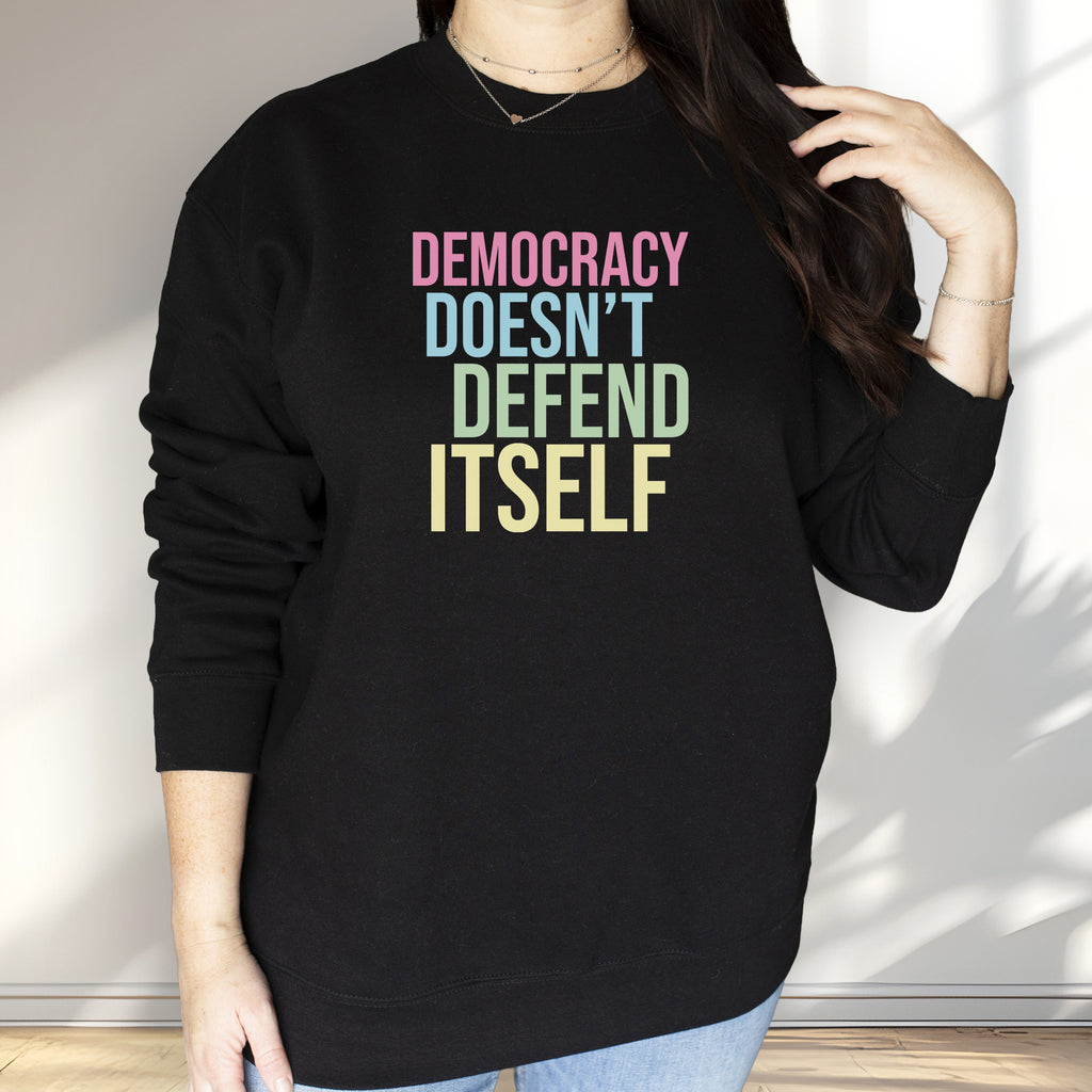 Female model wearing black sweatshirt that says “Democracy Doesn’t Defend Itself” in pink, blue, green, and yellow.