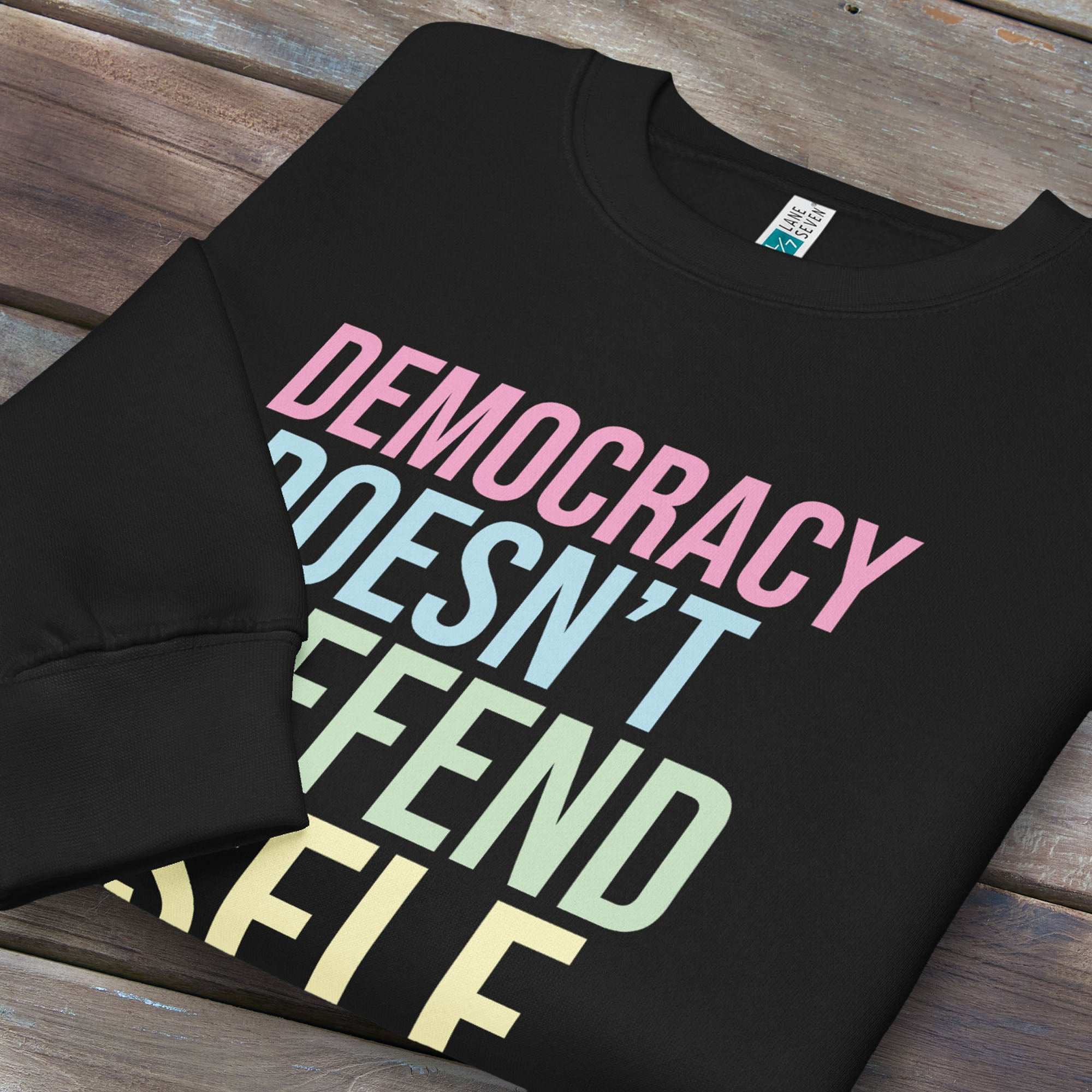Folded black sweatshirt that says “Democracy Doesn’t Defend Itself” in pink, blue, green, and yellow.