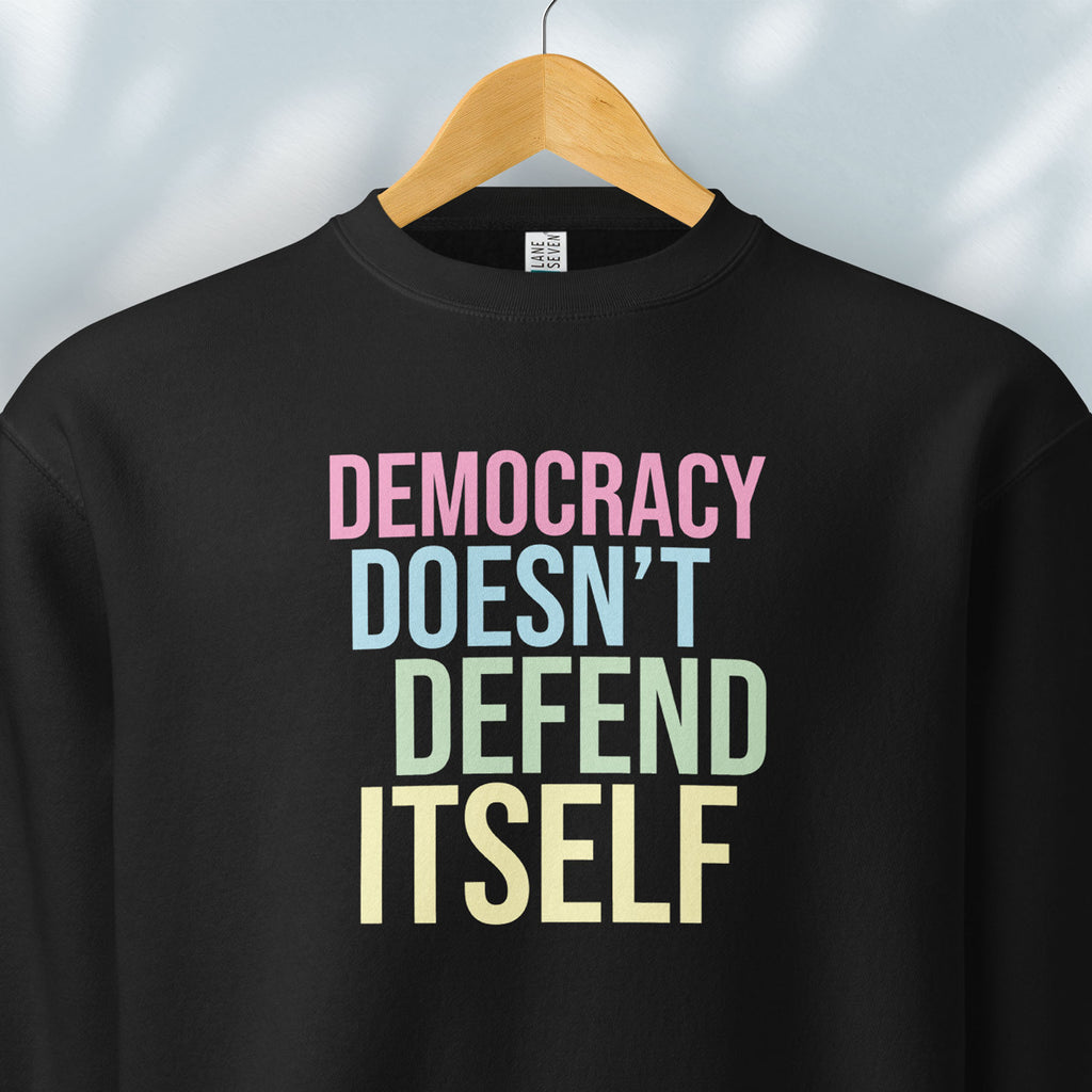 Hanging black sweatshirt that says “Democracy Doesn’t Defend Itself” in pink, blue, green, and yellow.