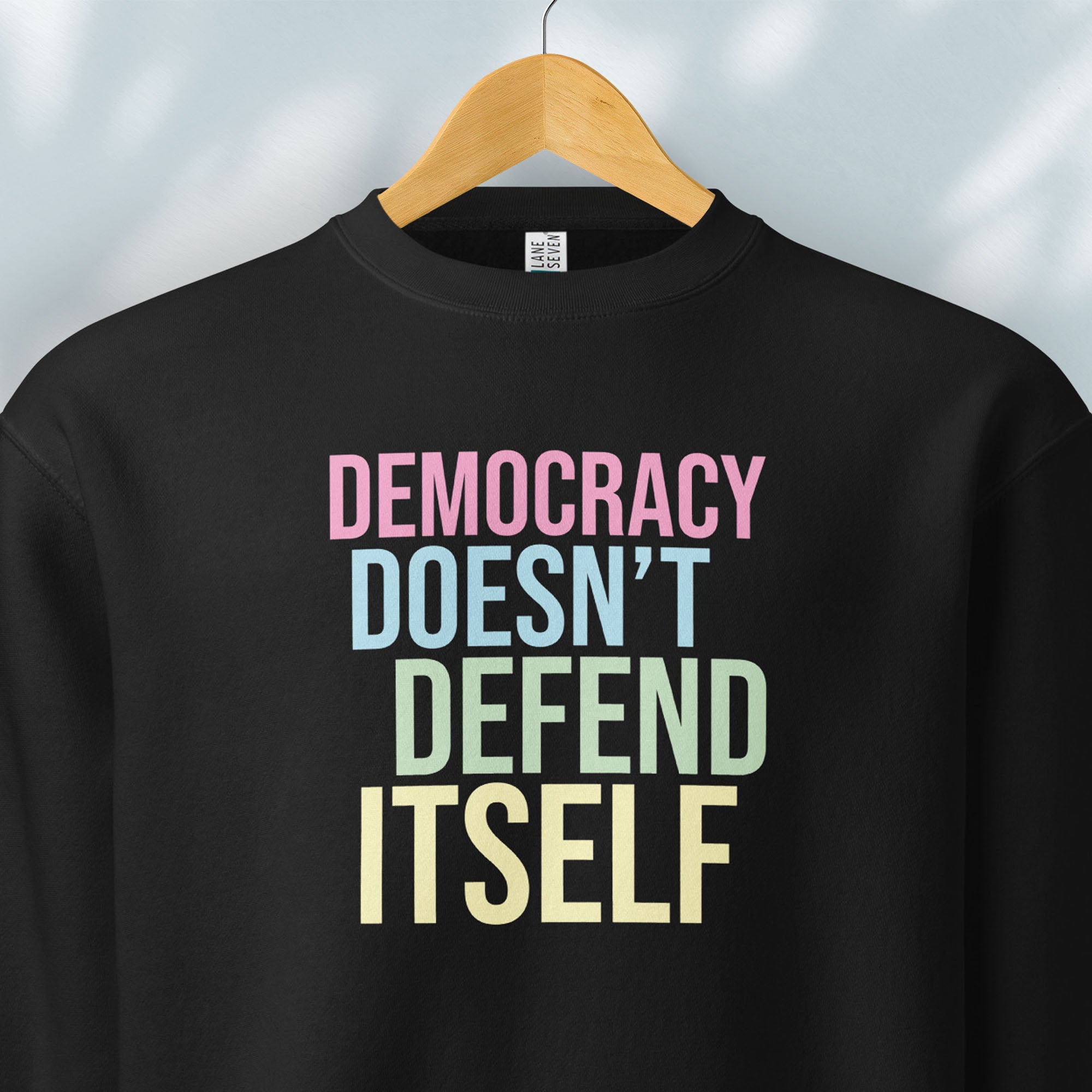 Hanging black sweatshirt that says “Democracy Doesn’t Defend Itself” in pink, blue, green, and yellow.
