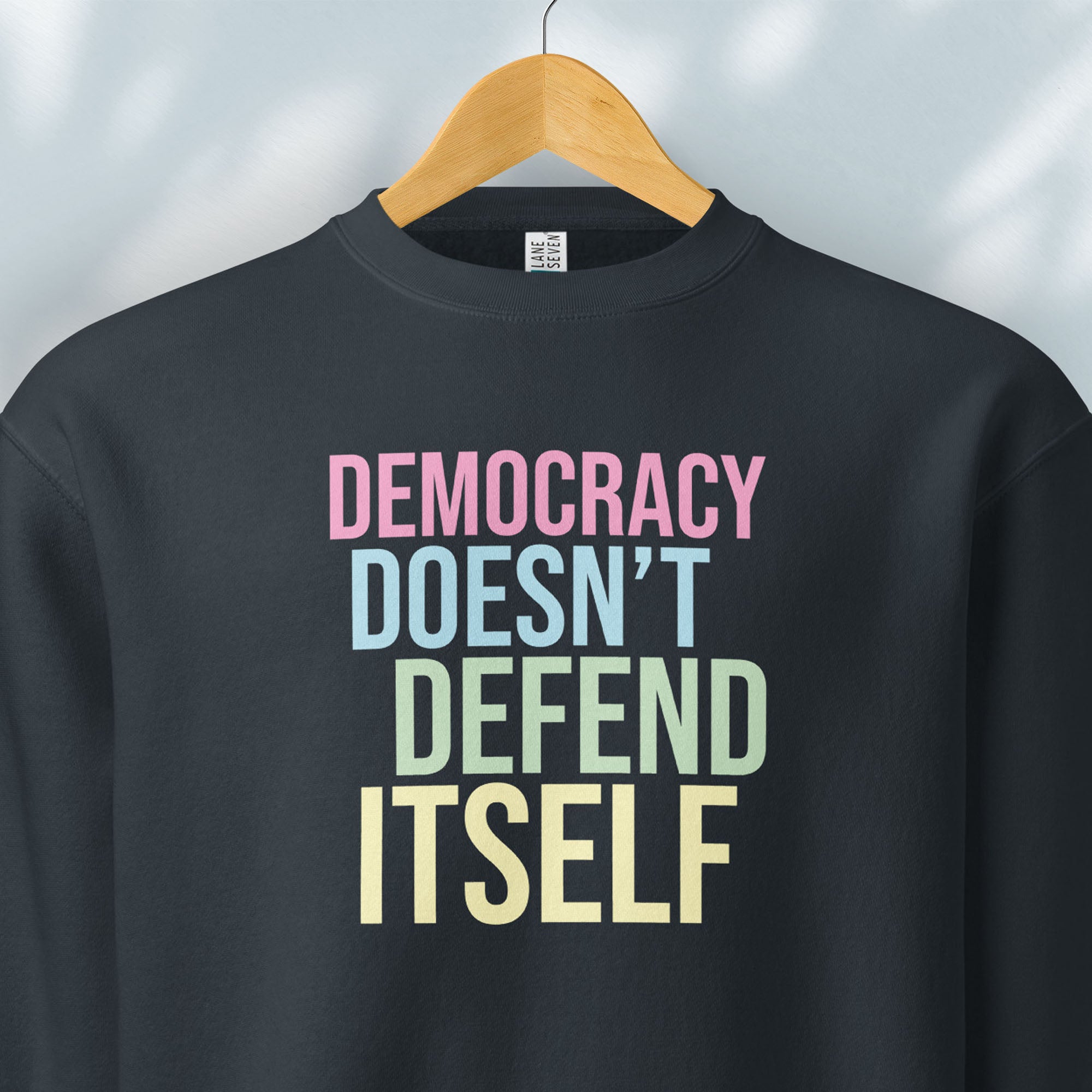 Hanging navy sweatshirt that says “Democracy Doesn’t Defend Itself” in pink, blue, green, and yellow.