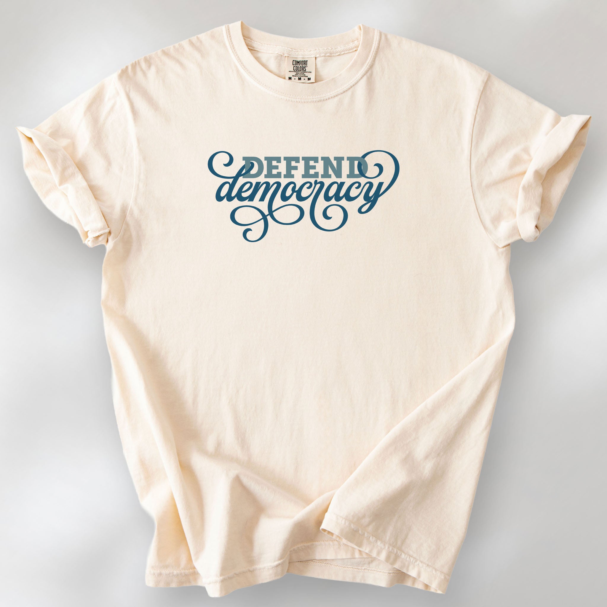 Ivory t-shirt with blue slab and script text reading “Defend Democracy.”