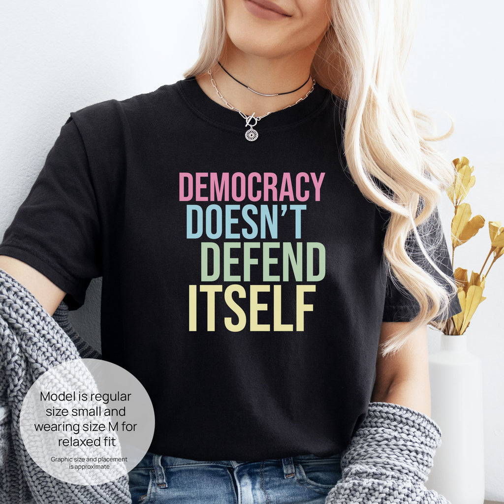 Female model wearing black unisex political t-shirt that says “Democracy Doesn’t Defend Itself” in pink, blue, green, and yellow on a black Comfort Colors tee. Includes size and fit information for the model who is regular size small, and wearing a size M shirt for a relaxed fit.
