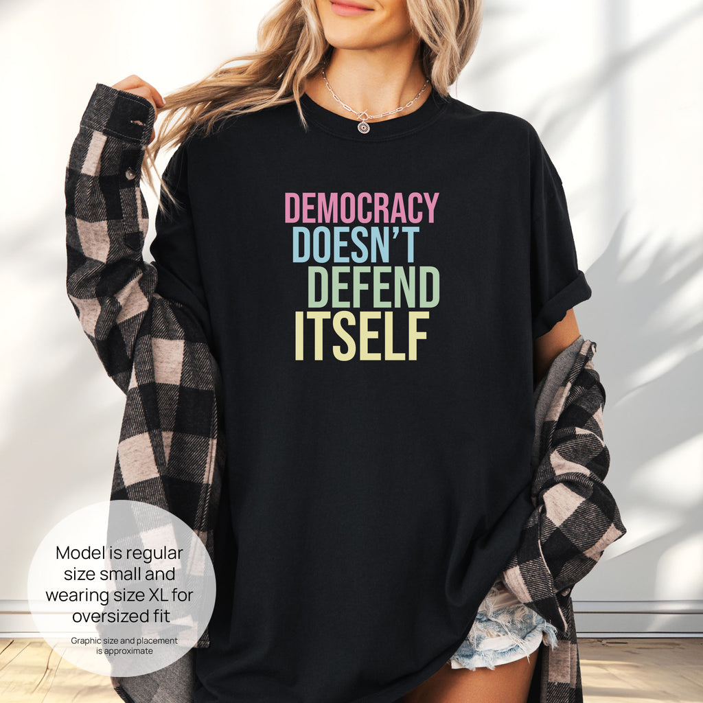 Female model wearing oversized black unisex political t-shirt that says “Democracy Doesn’t Defend Itself” in pink, blue, green, and yellow on a black Comfort Colors tee. Includes size and fit information for the model who is regular size small, and wearing a size XL shirt for an oversized fit.
