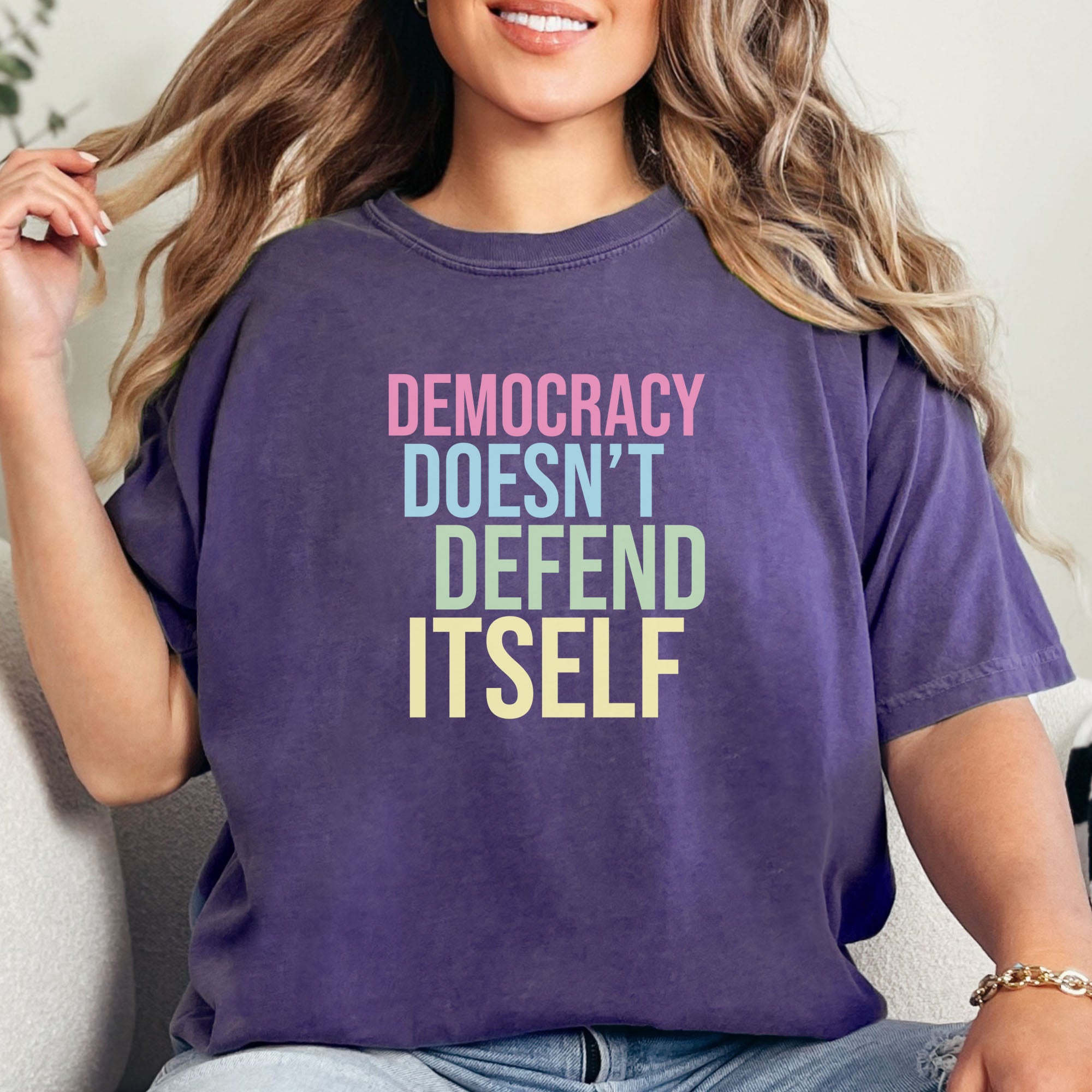 Female model wearing grape unisex political t-shirt that says “Democracy Doesn’t Defend Itself” in pink, blue, green, and yellow on a black Comfort Colors 1717 shirt.