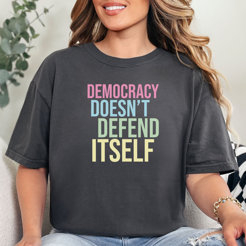 Female model wearing graphite unisex political t-shirt that says “Democracy Doesn’t Defend Itself” in pink, blue, green, and yellow on a black Comfort Colors 1717 shirt.