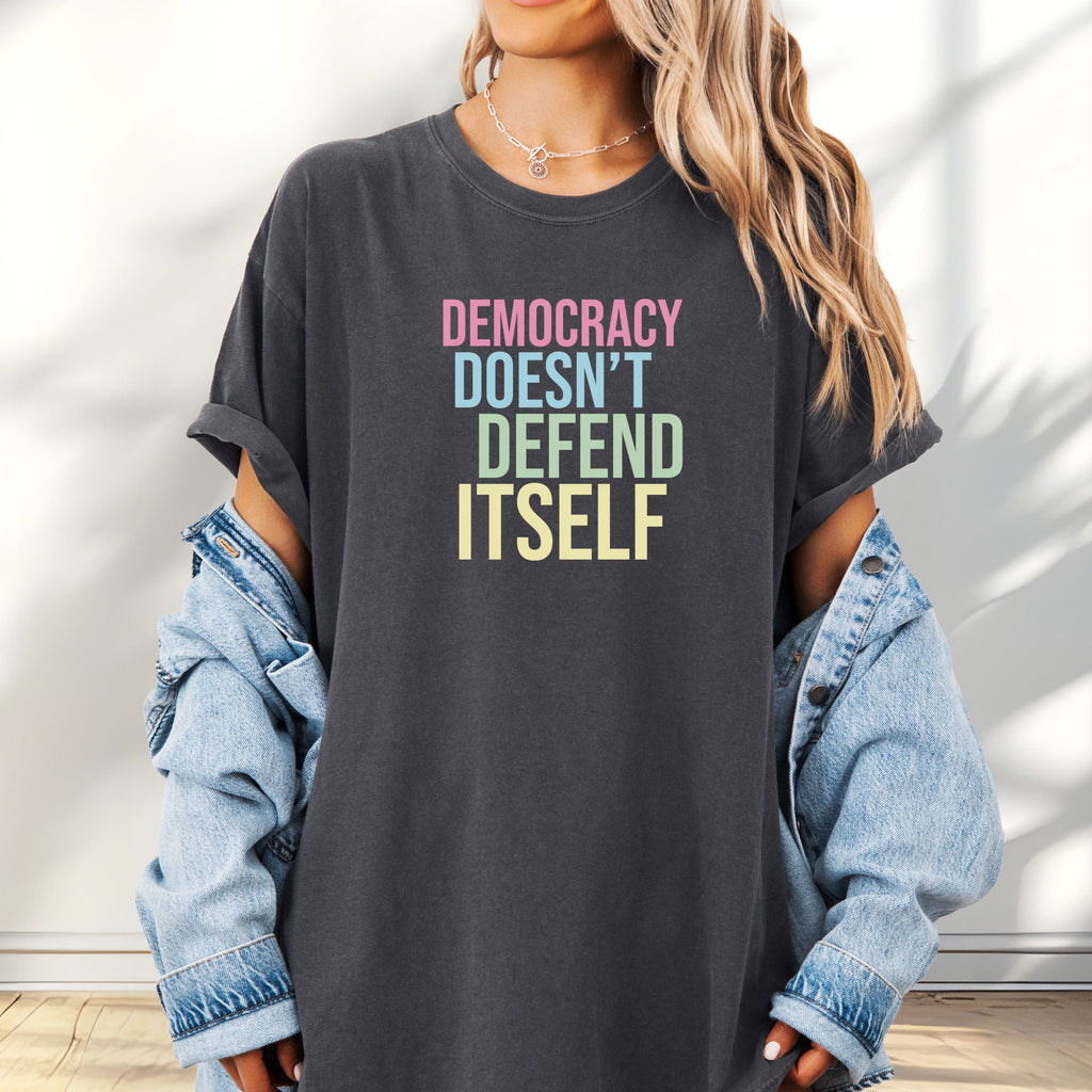Female model wearing oversized pepper unisex political t-shirt that says “Democracy Doesn’t Defend Itself” in pink, blue, green, and yellow on a black Comfort Colors tee.
