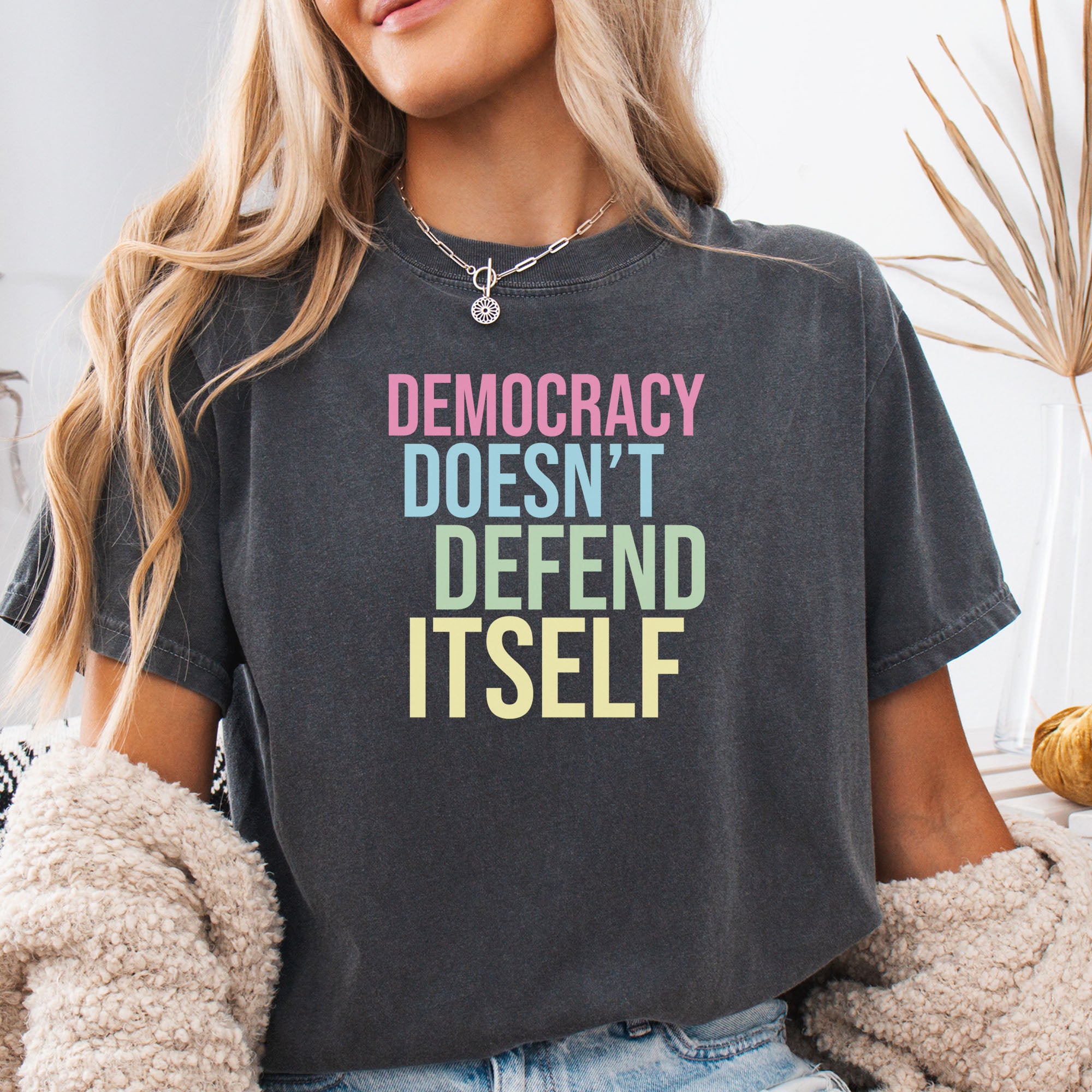 Female model wearing pepper unisex political t-shirt that says “Democracy Doesn’t Defend Itself” in pink, blue, green, and yellow on a black Comfort Colors tee.