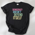 Black unisex political t-shirt that says “Democracy Doesn’t Defend Itself” in pink, blue, green, and yellow on a black Comfort Colors 1717 shirt.