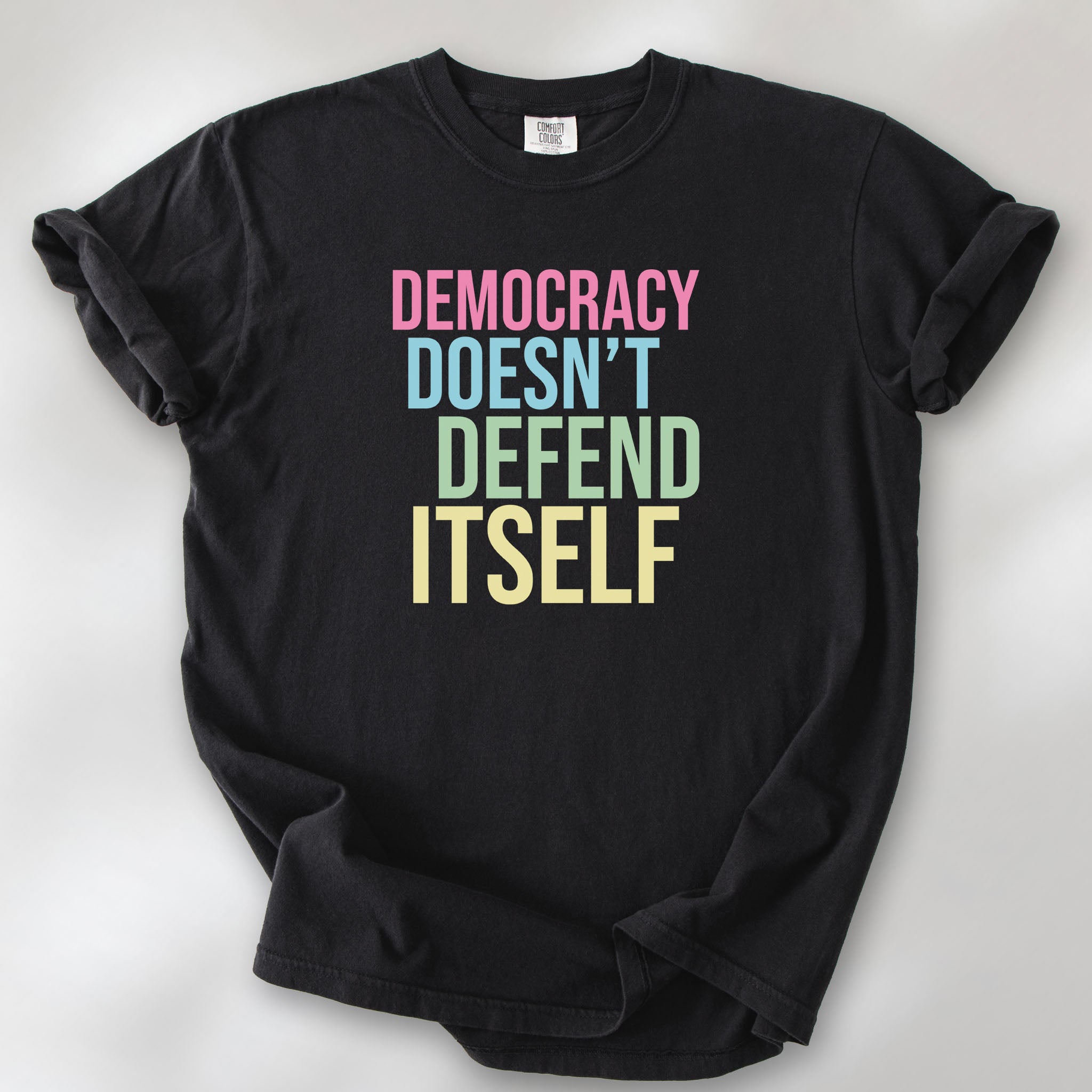 Black unisex political t-shirt that says “Democracy Doesn’t Defend Itself” in pink, blue, green, and yellow on a black Comfort Colors 1717 shirt.