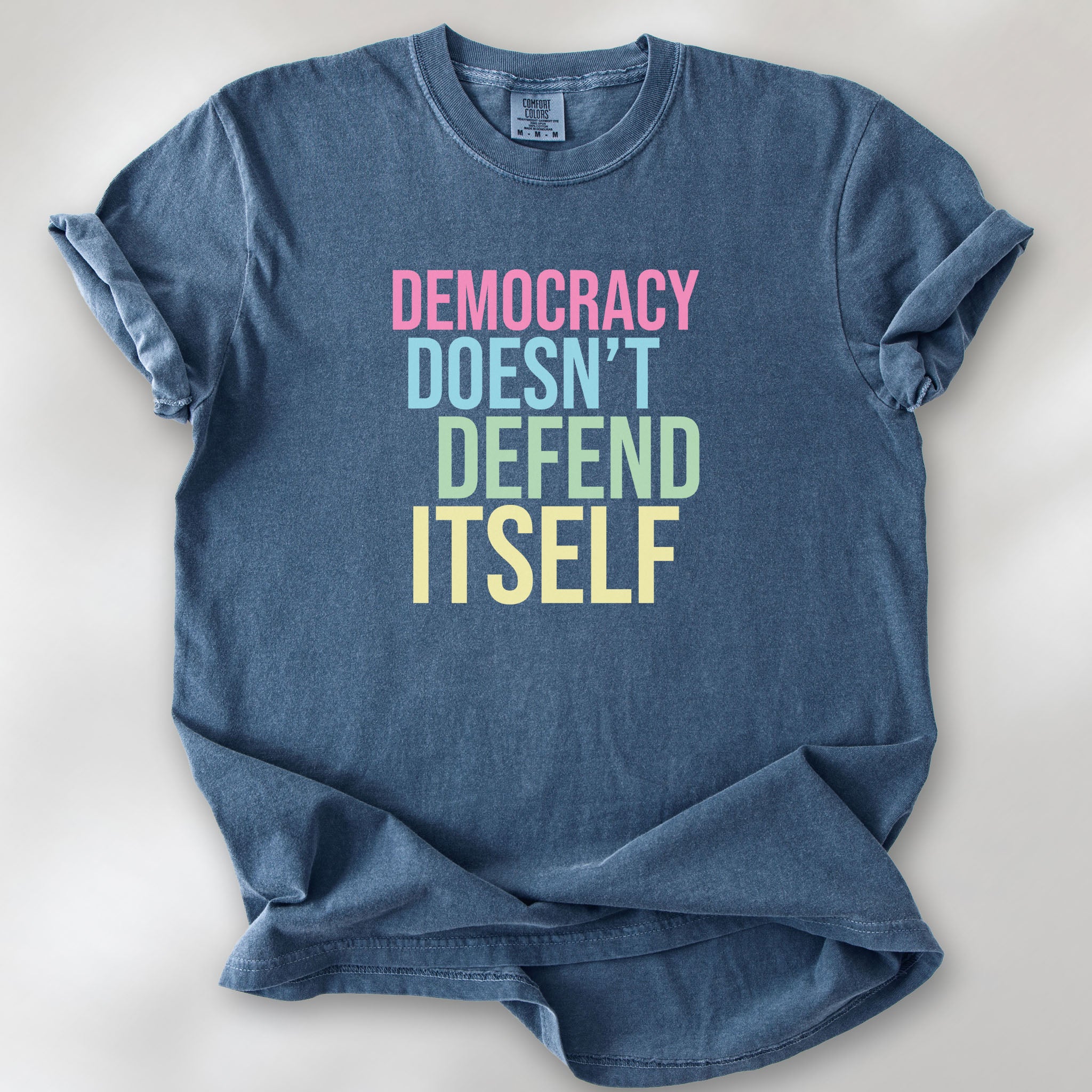 Denim unisex political t-shirt that says “Democracy Doesn’t Defend Itself” in pink, blue, green, and yellow on a black Comfort Colors 1717 shirt.
