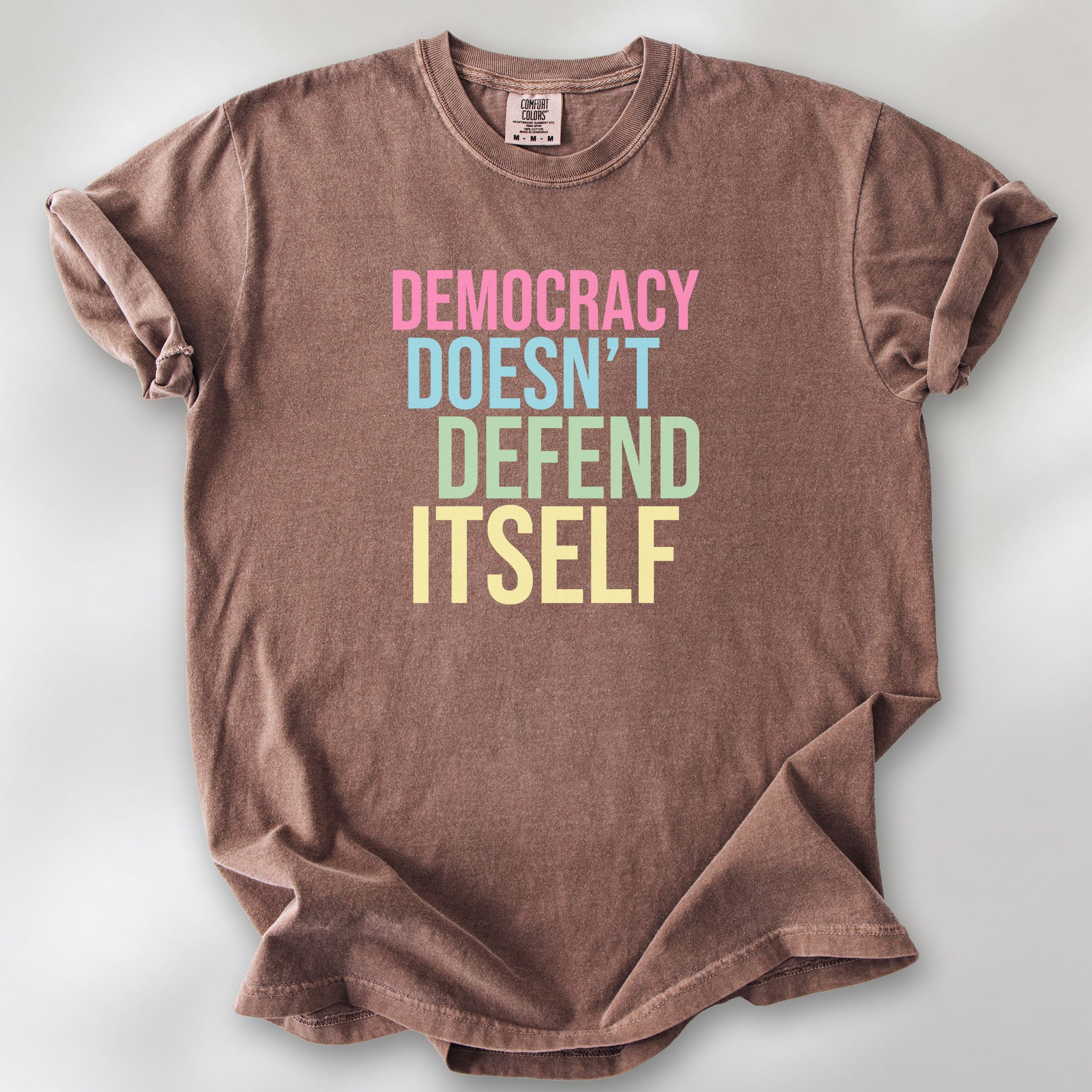 Espresso unisex political t-shirt that says “Democracy Doesn’t Defend Itself” in pink, blue, green, and yellow on a black Comfort Colors 1717 shirt.