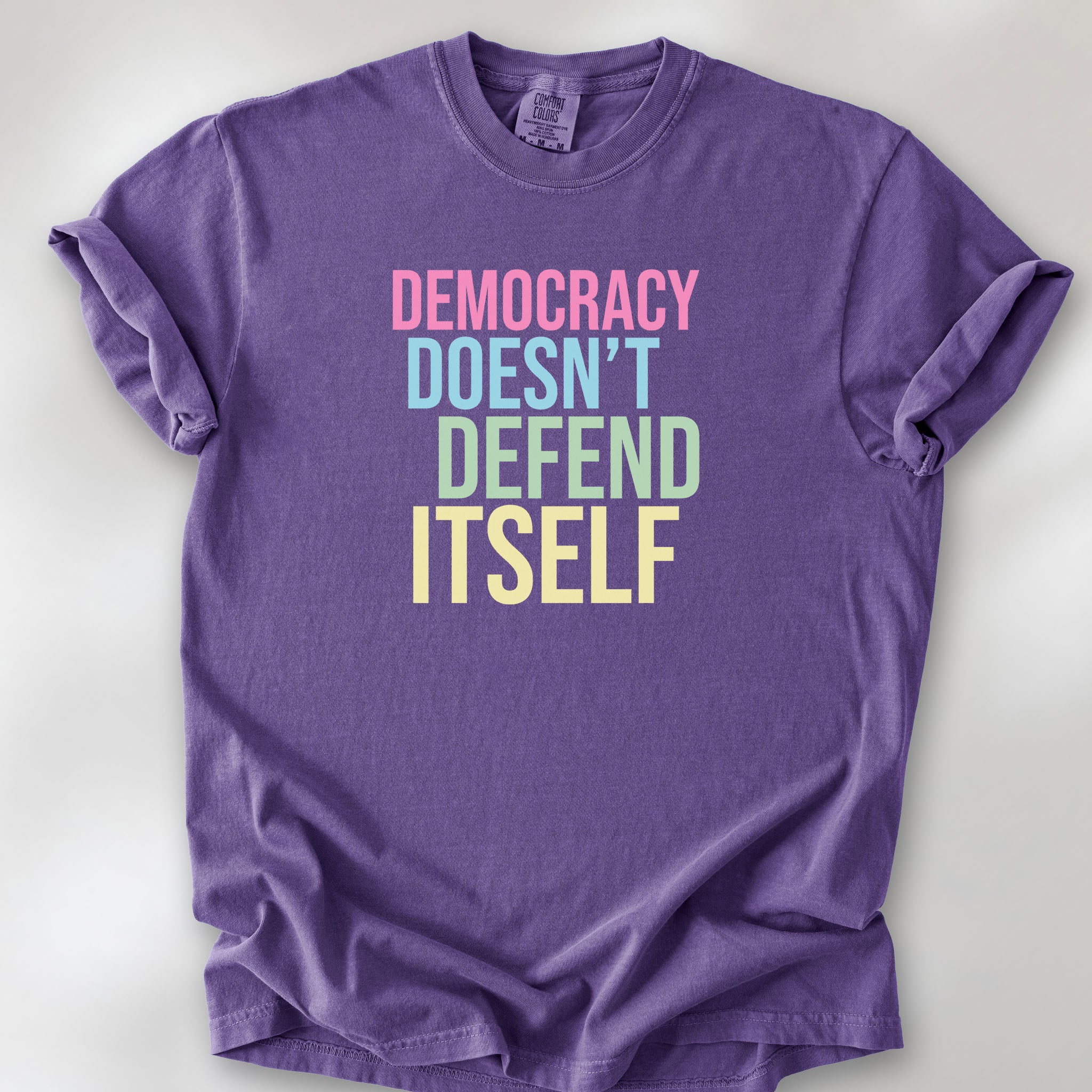 Grape unisex political t-shirt that says “Democracy Doesn’t Defend Itself” in pink, blue, green, and yellow on a black Comfort Colors 1717 shirt.