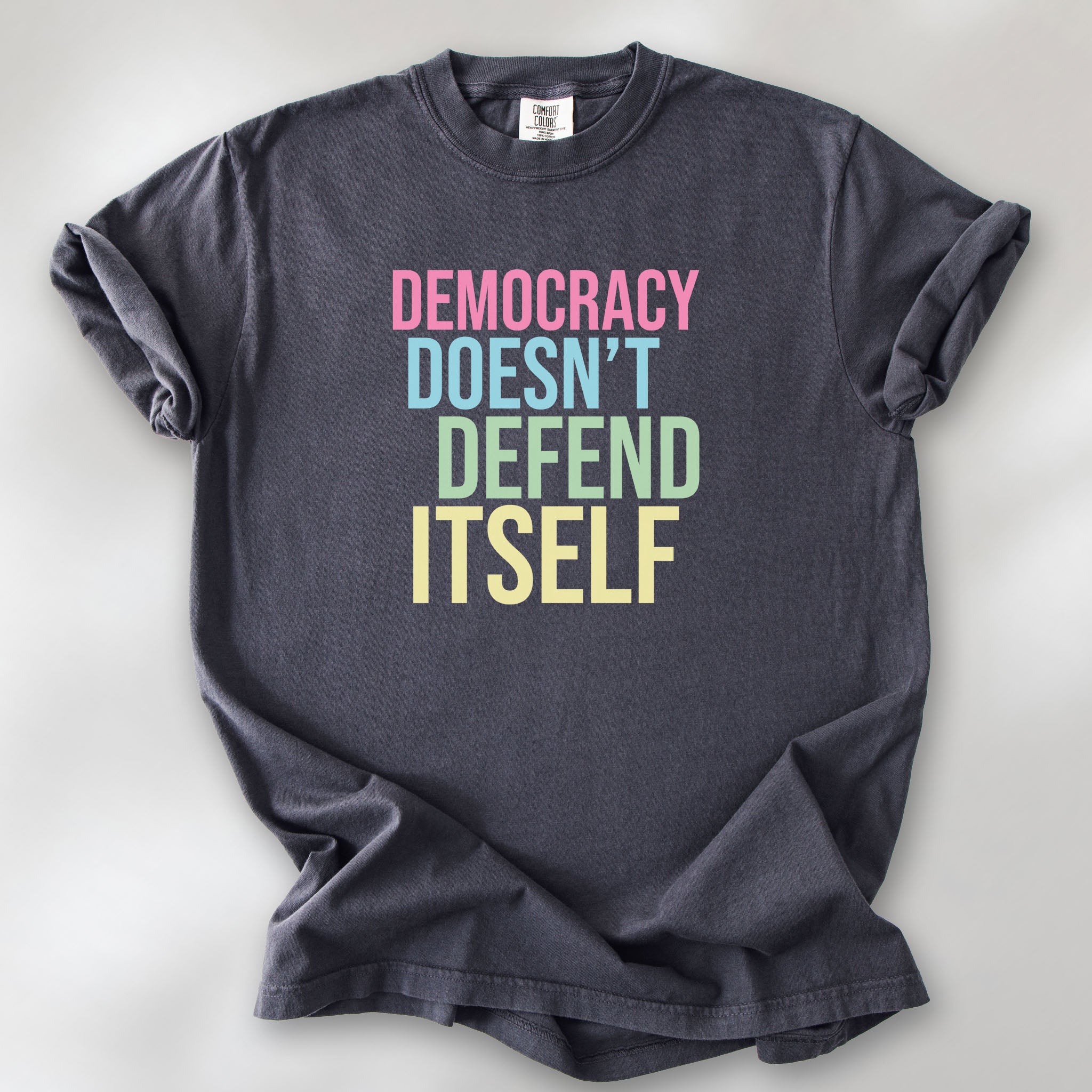 Graphite unisex political t-shirt that says “Democracy Doesn’t Defend Itself” in pink, blue, green, and yellow on a black Comfort Colors 1717 shirt.