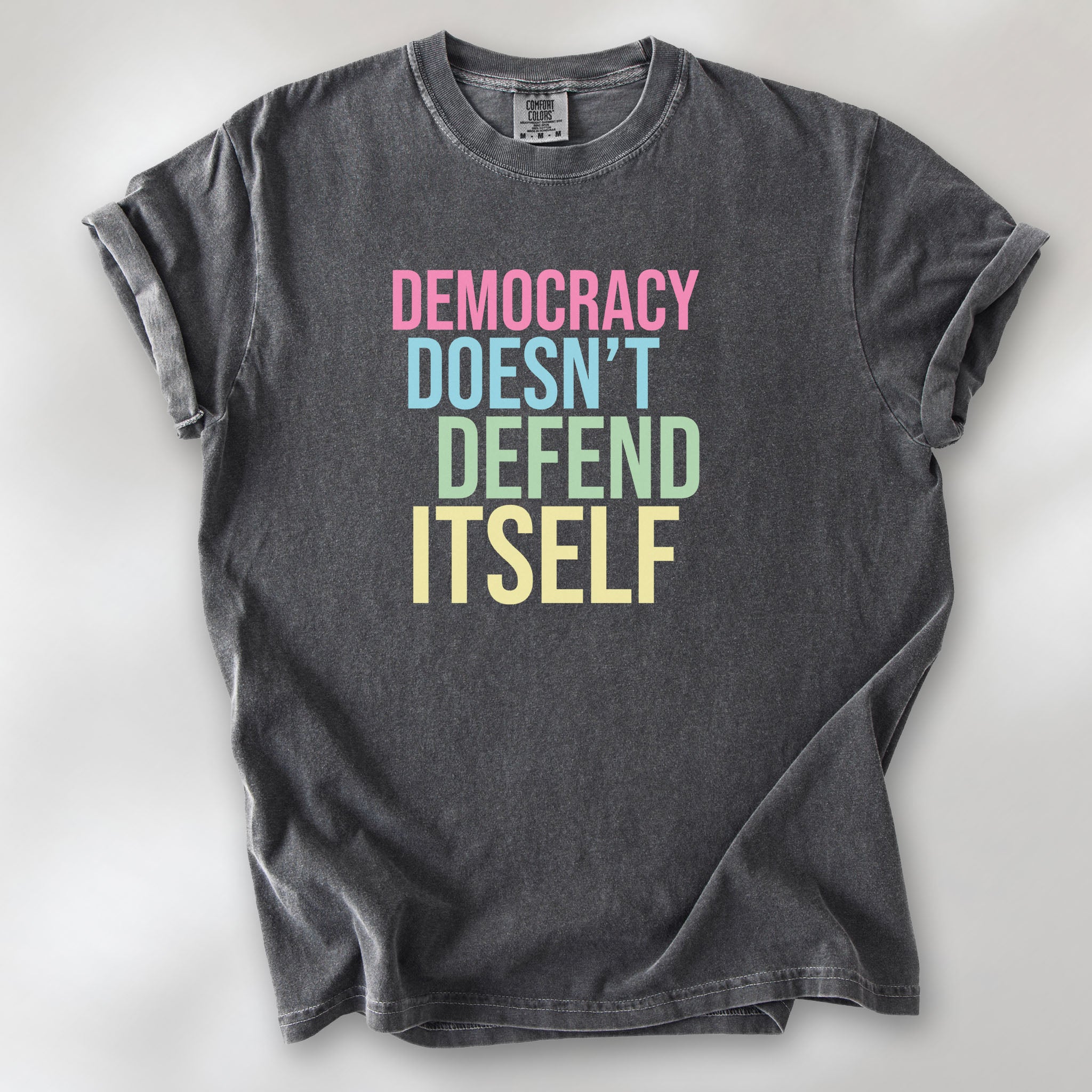 Pepper unisex political t-shirt that says “Democracy Doesn’t Defend Itself” in pink, blue, green, and yellow on a black Comfort Colors 1717 shirt.