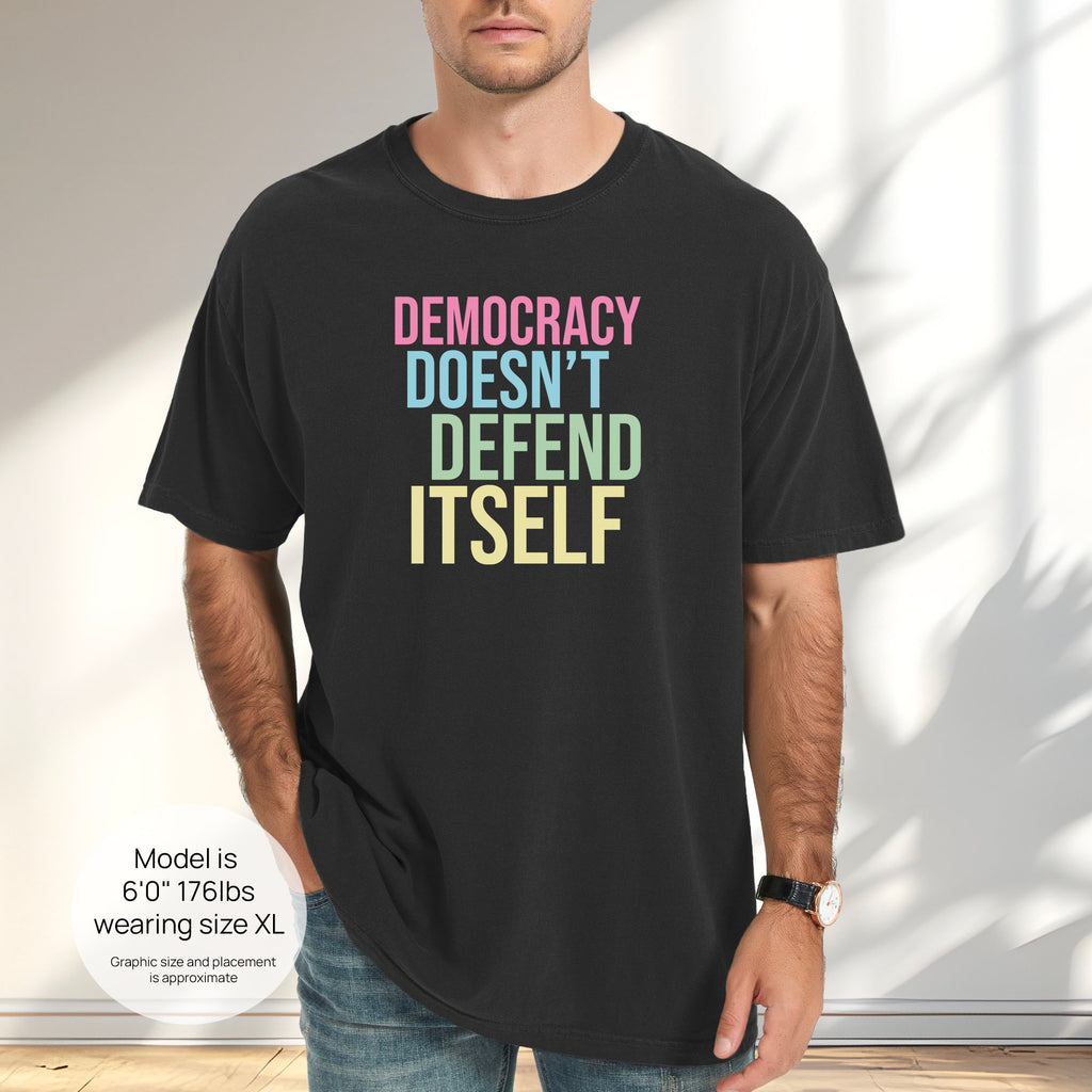 Male model wearing black unisex political t-shirt that says “Democracy Doesn’t Defend Itself” in pink, blue, green, and yellow on a black Comfort Colors tee. Includes size and fit information for the model who is 6 feet 0 inches, 176 pounds, and wearing a size XL shirt.
