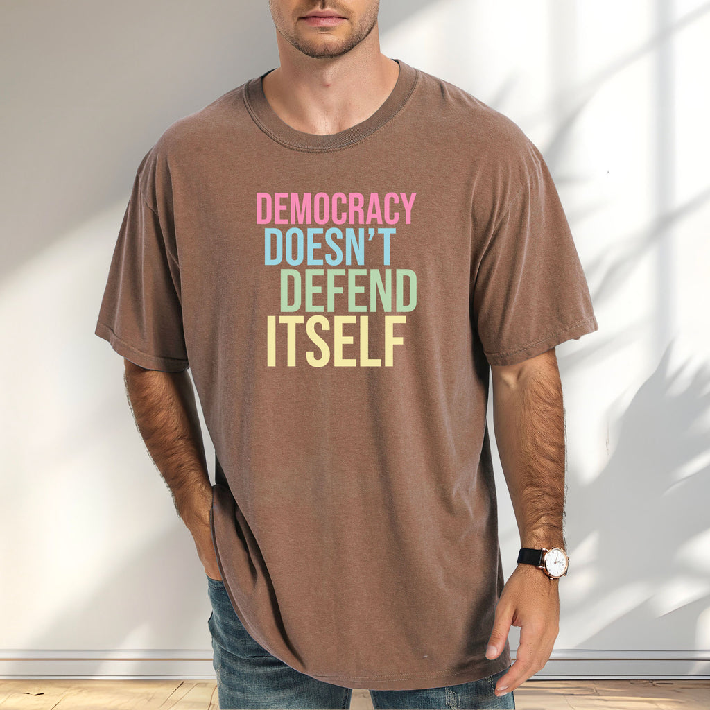 Male model wearing espresso unisex political t-shirt that says “Democracy Doesn’t Defend Itself” in pink, blue, green, and yellow on a black Comfort Colors 1717 shirt.