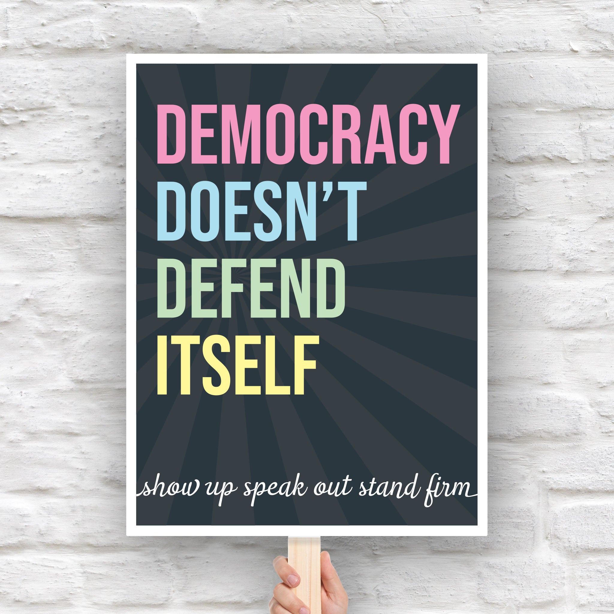 Downloadable, printable protest sign with dark background with subtle burst, bold colorful text that reads "democracy doesn't defend itself", then white script text near the bottom that reads "show up, speak out, stand firm."