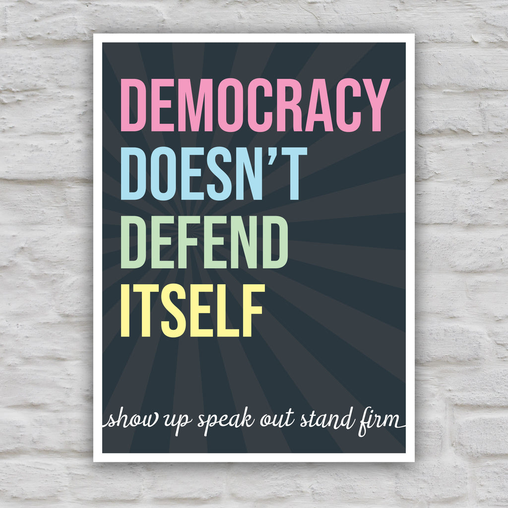 Color version of protest sign with dark background with subtle burst, bold colorful text that reads "democracy doesn't defend itself", then white script text near the bottom that reads "show up, speak out, stand firm."