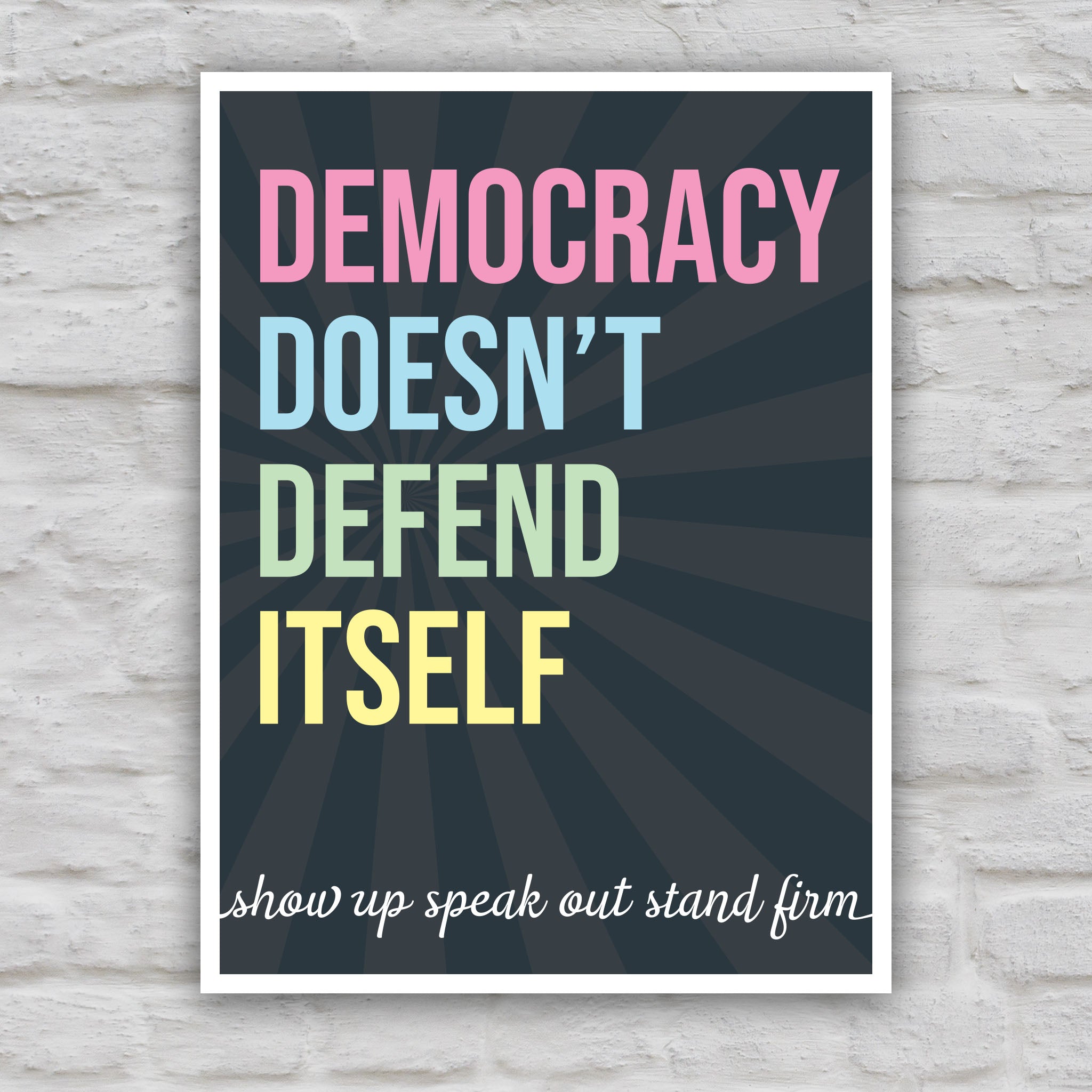 Color version of protest sign with dark background with subtle burst, bold colorful text that reads "democracy doesn't defend itself", then white script text near the bottom that reads "show up, speak out, stand firm."