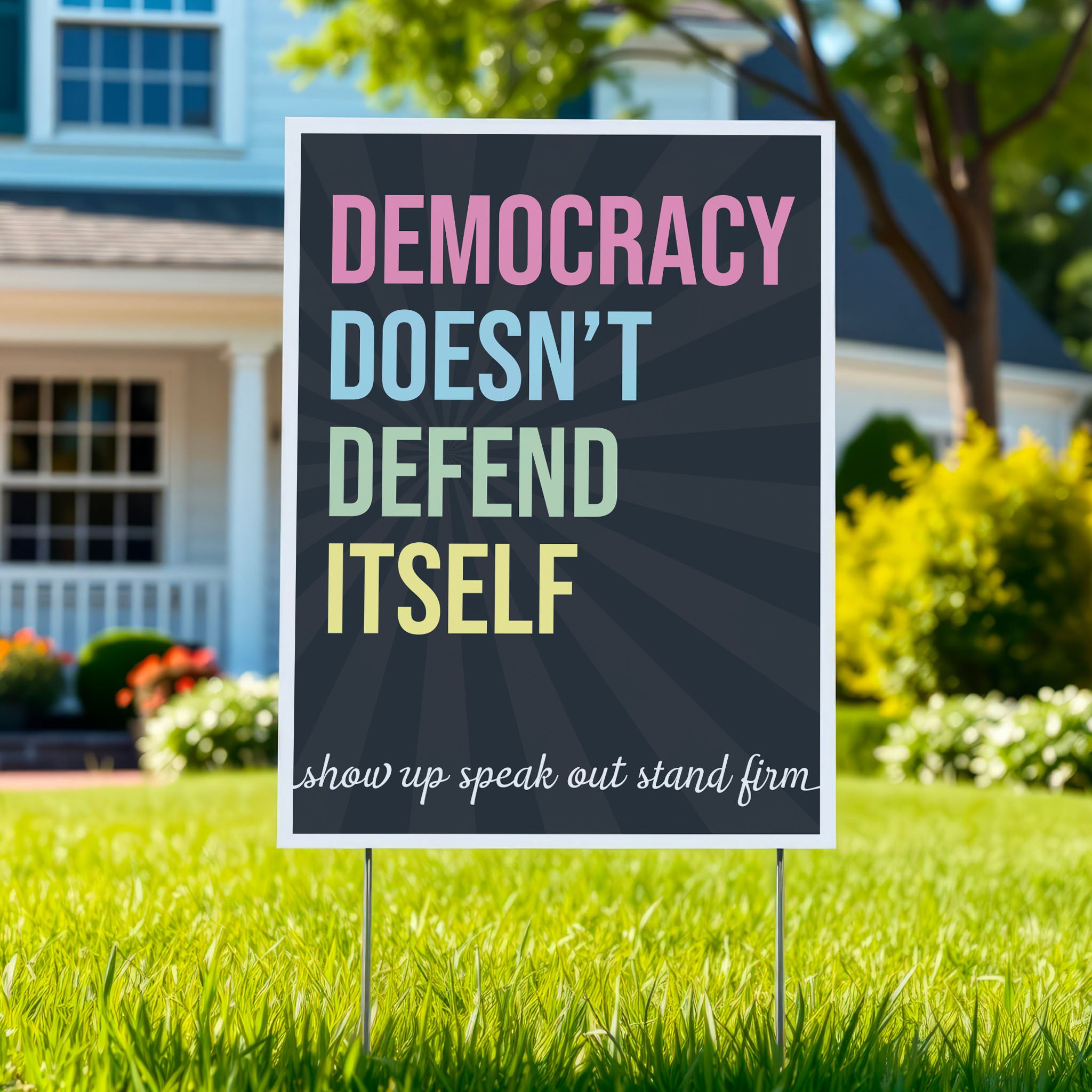 Yard sign with dark background with subtle burst, bold colorful text that reads "democracy doesn't defend itself", then white script text near the bottom that reads "show up, speak out, stand firm."