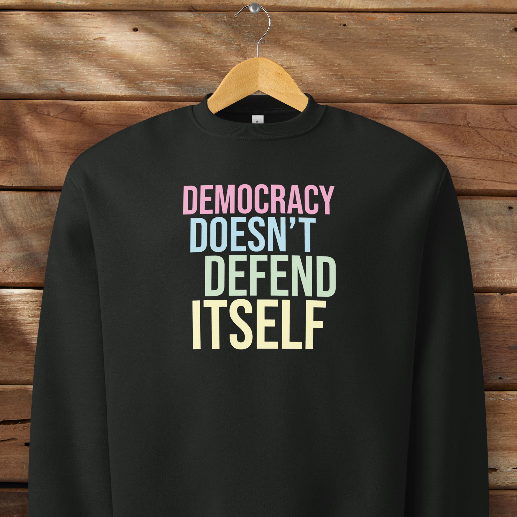 Black unisex crewneck sweatshirt with bright bold text that reads "democracy doesn't defend itself", on a hanger.