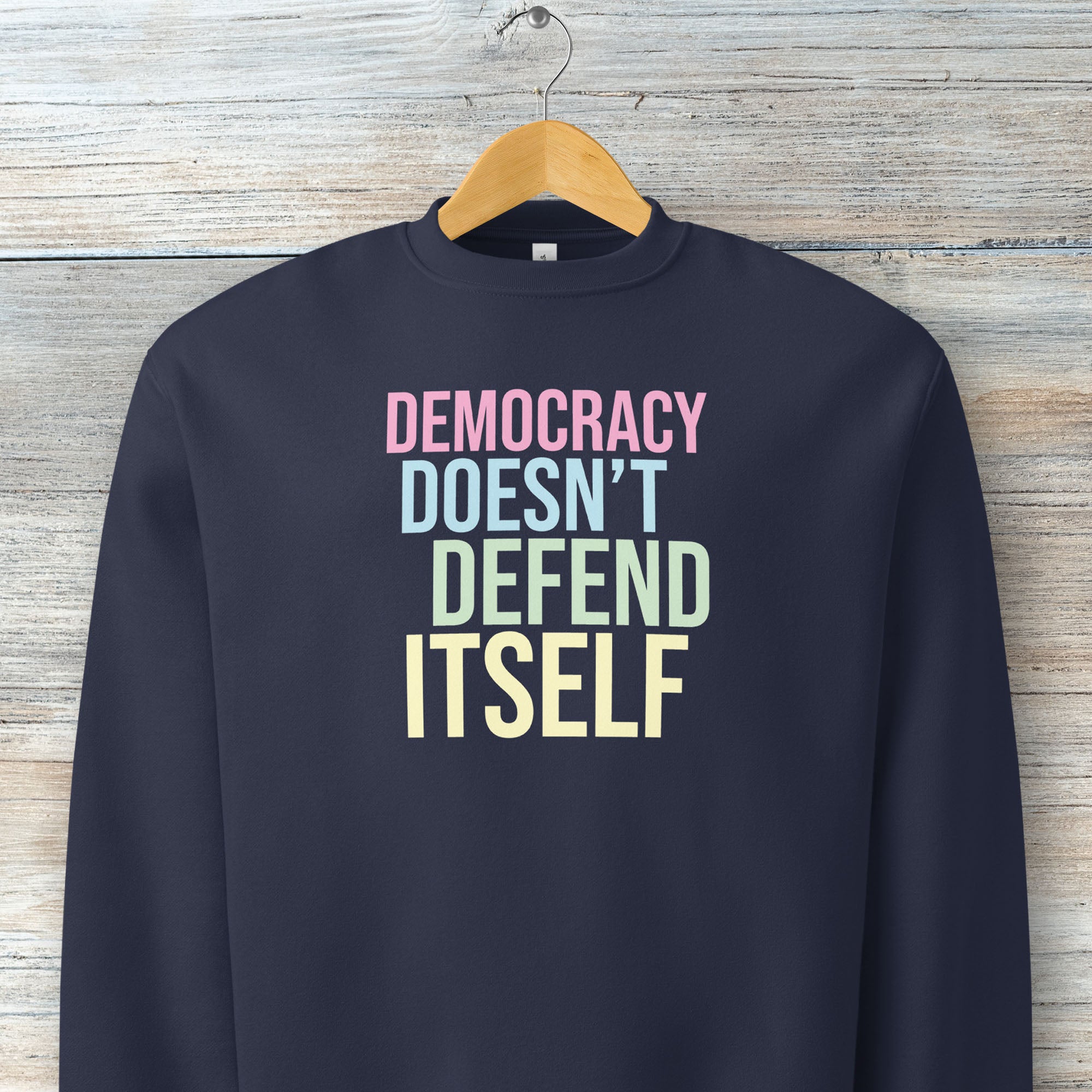 Navy unisex crewneck sweatshirt with bright bold text that reads "democracy doesn't defend itself", on a hanger.