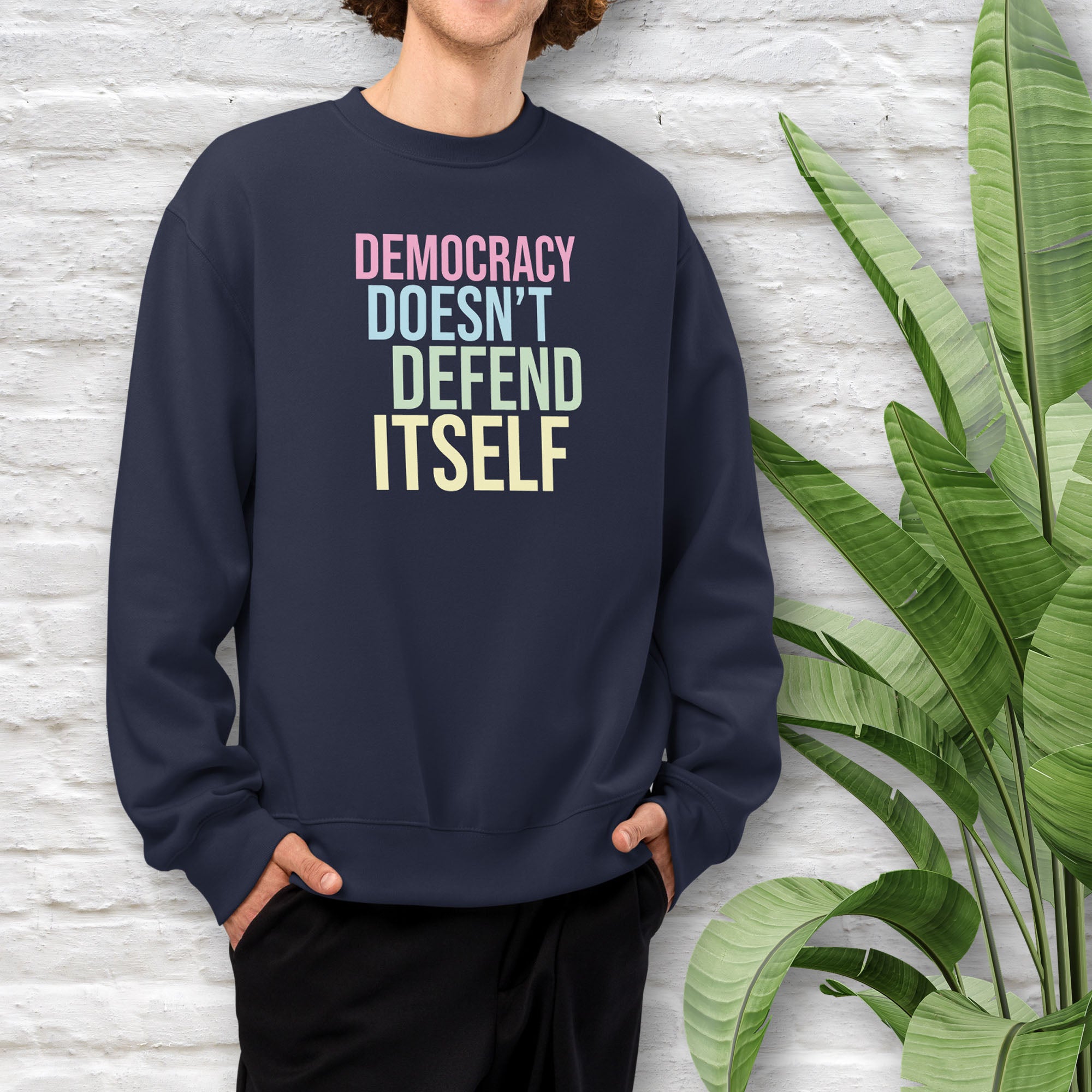 Male model wearing navy unisex crewneck sweatshirt with bright bold text that reads "democracy doesn't defend itself".