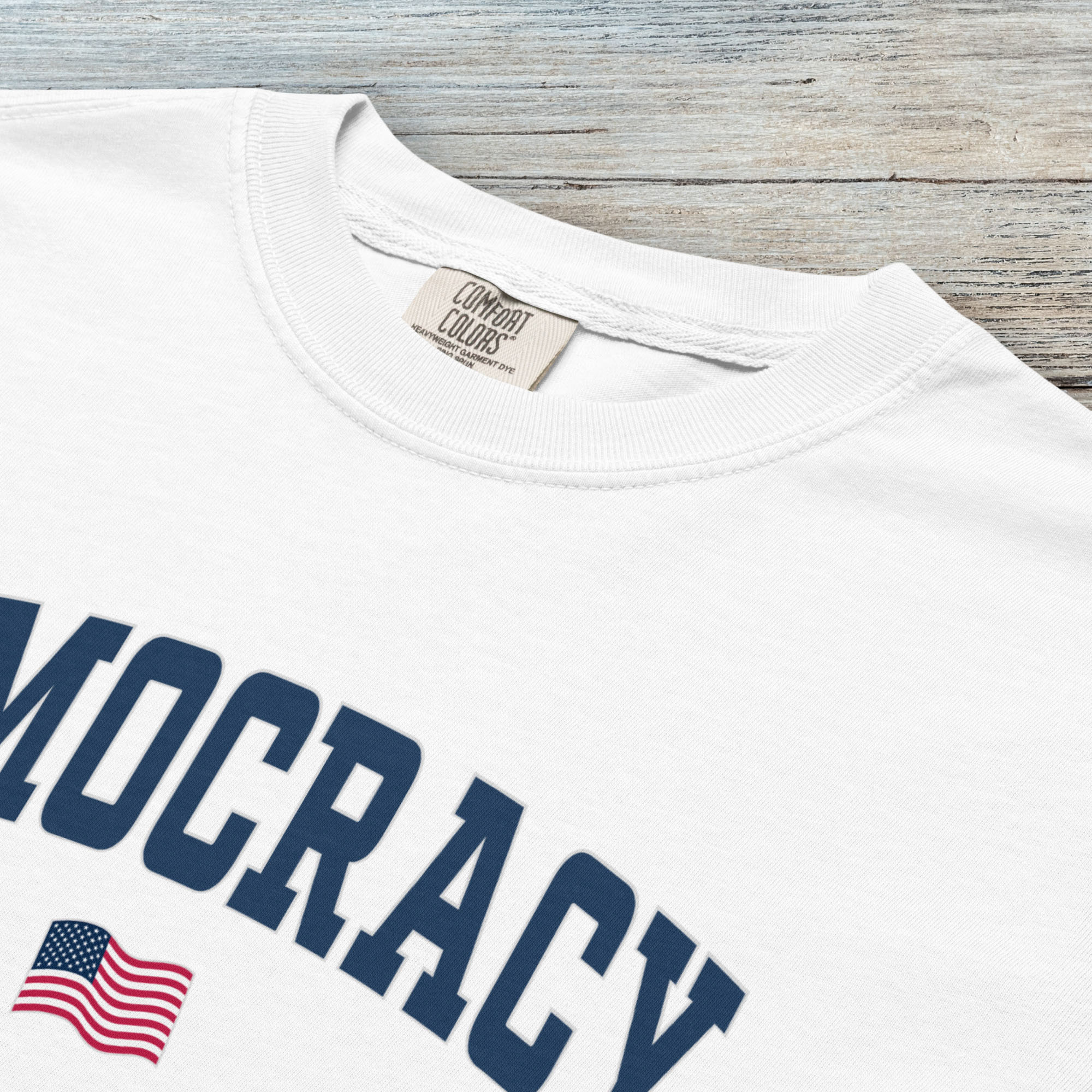 Closeup detail of white t-shirt with the word “Democracy” in bold navy letters and a small American flag underneath, symbolizing U.S. pride in democracy over authoritarianism, tyranny, dictators.