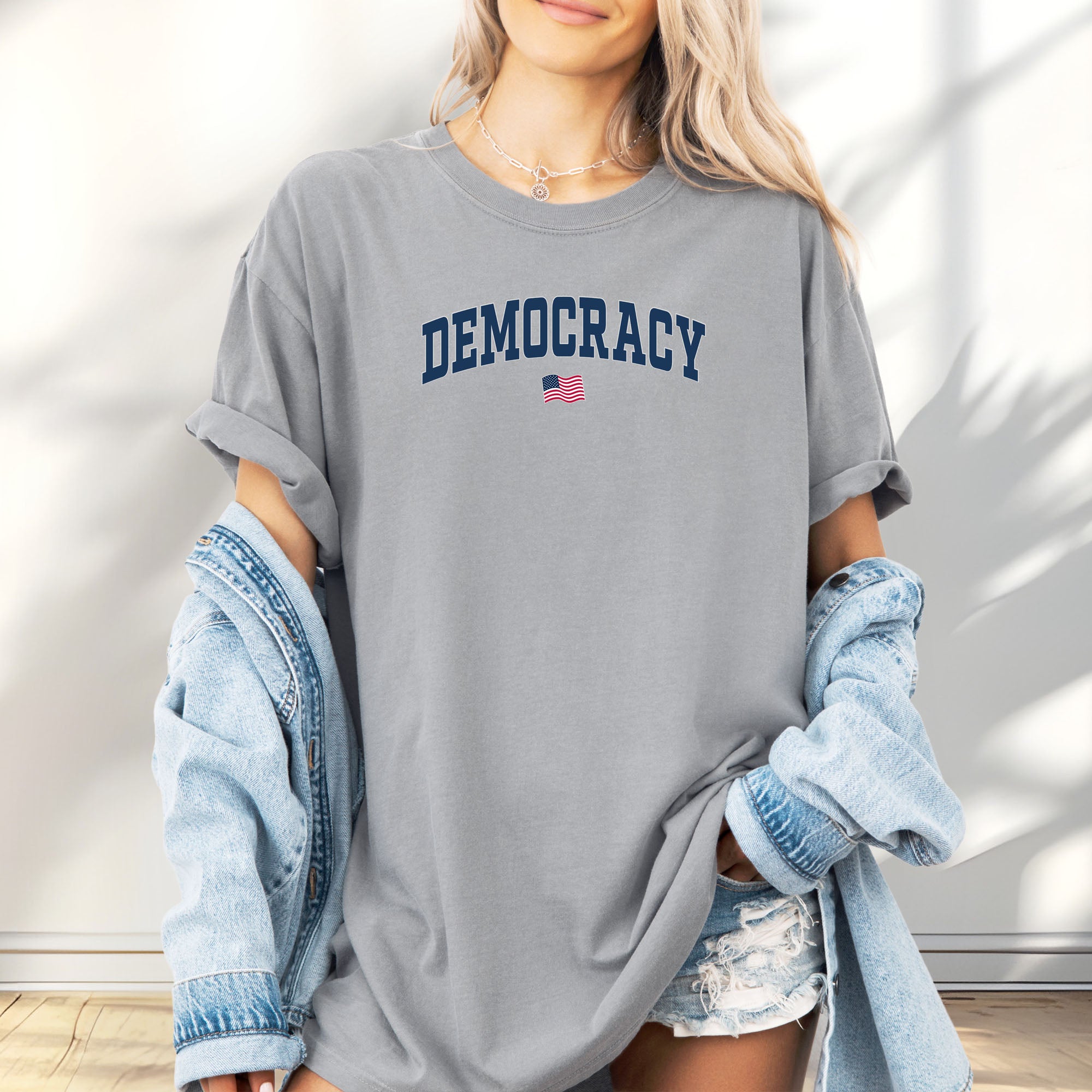 Female model wearing granite t-shirt with the word “Democracy” in bold navy letters and a small American flag underneath, symbolizing U.S. pride in democracy over authoritarianism, tyranny, dictators.