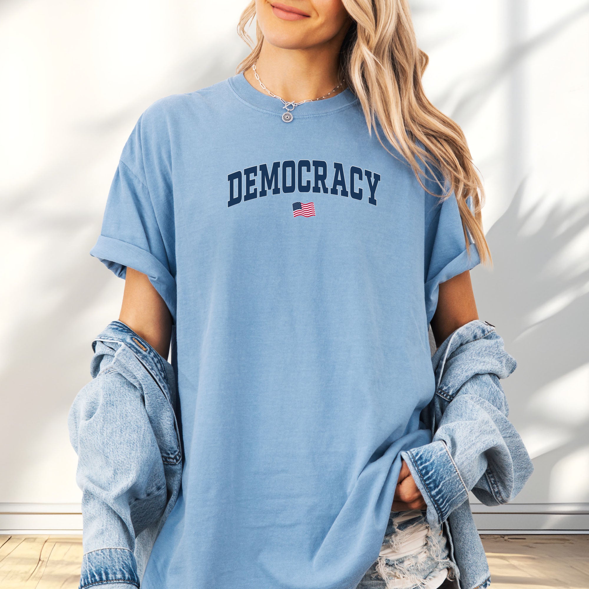 Female model wearing oversized washed denim t-shirt with the word “Democracy” in bold navy letters and a small American flag underneath, symbolizing U.S. pride in democracy over authoritarianism, tyranny, dictators.