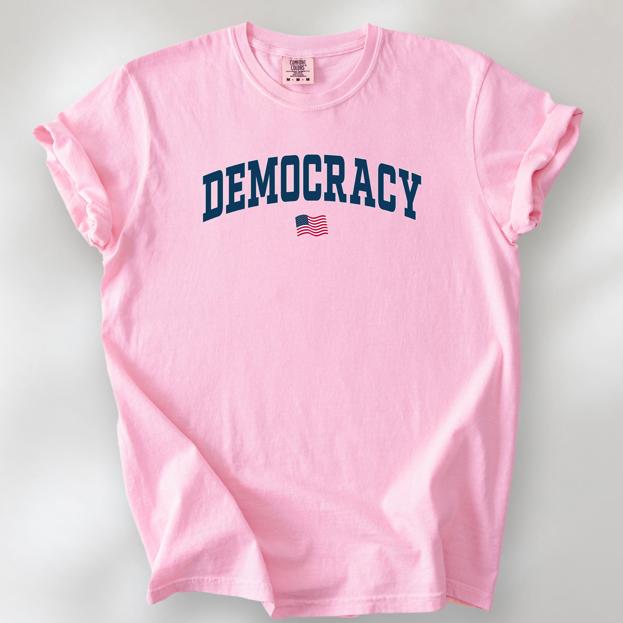 Blossom t-shirt with the word “Democracy” in bold navy letters and a small American flag underneath, symbolizing U.S. pride in democracy over authoritarianism, tyranny, dictators.