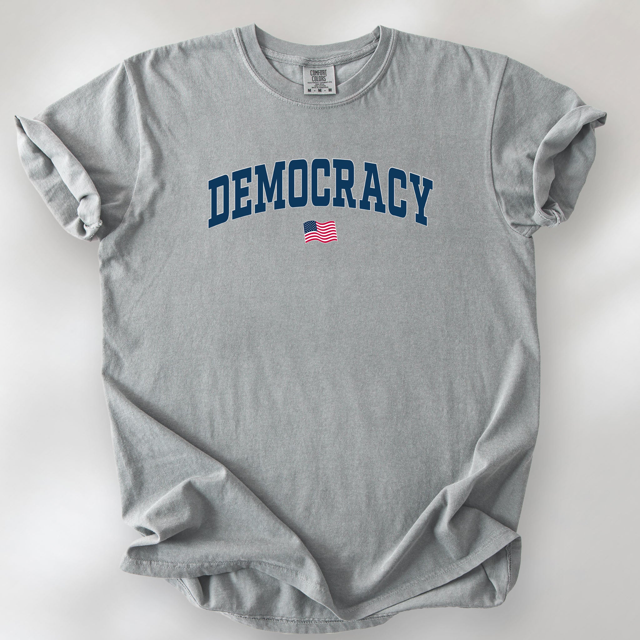Granite t-shirt with the word “Democracy” in bold navy letters and a small American flag underneath, symbolizing U.S. pride in democracy over authoritarianism, tyranny, dictators.