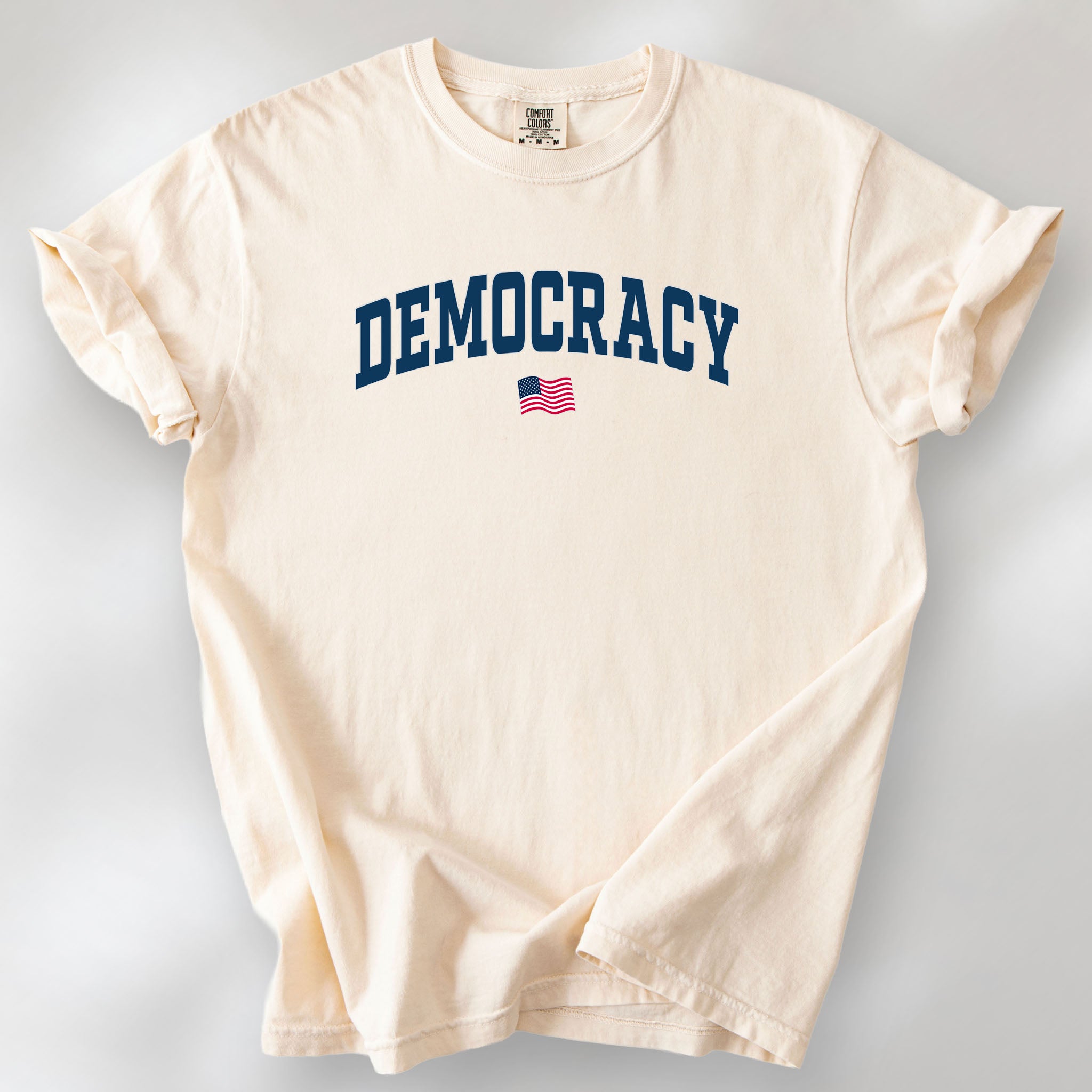 Ivory t-shirt with the word “Democracy” in bold navy letters and a small American flag underneath, symbolizing U.S. pride in democracy over authoritarianism, tyranny, dictators.
