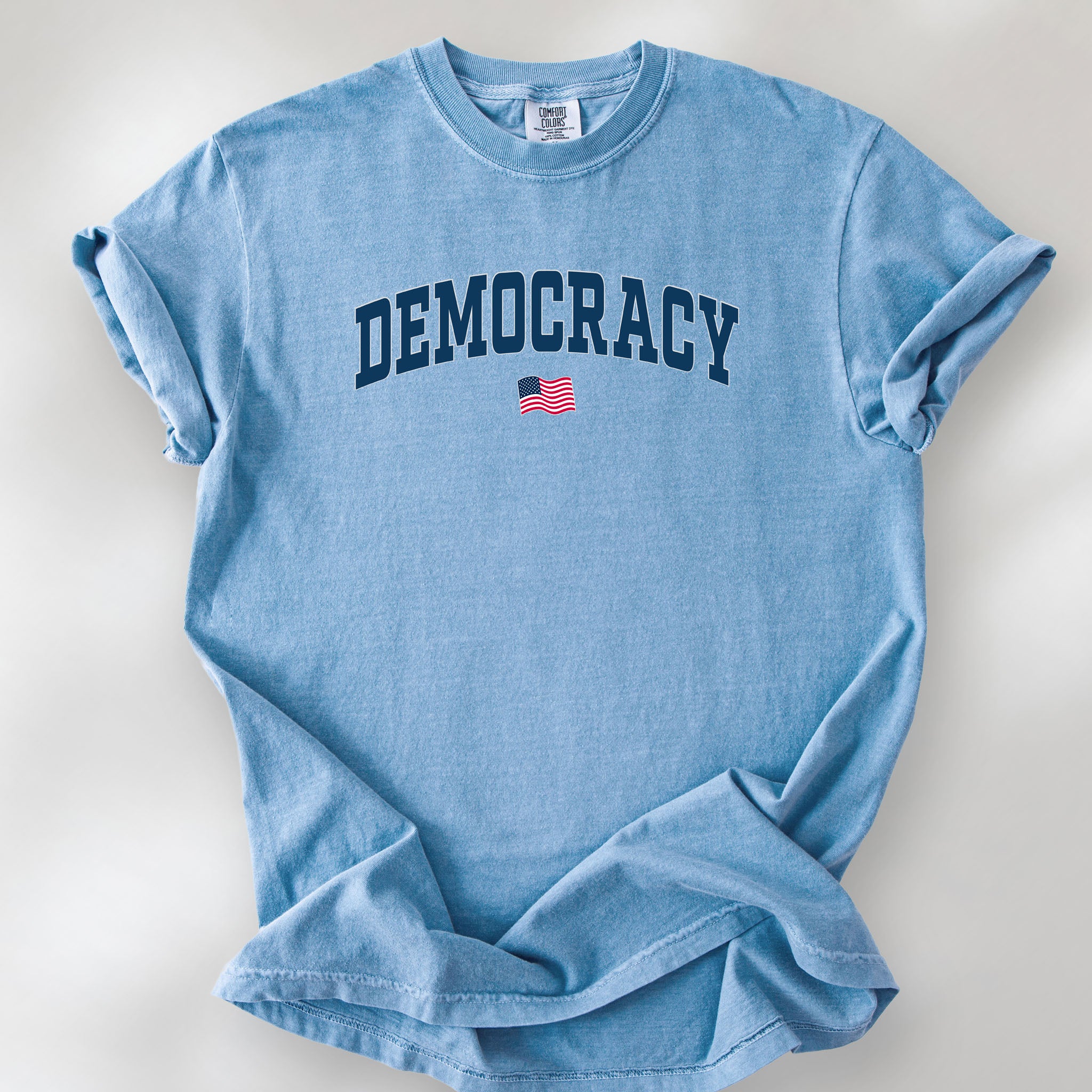 Washed denim t-shirt with the word “Democracy” in bold navy letters and a small American flag underneath, symbolizing U.S. pride in democracy over authoritarianism, tyranny, dictators.