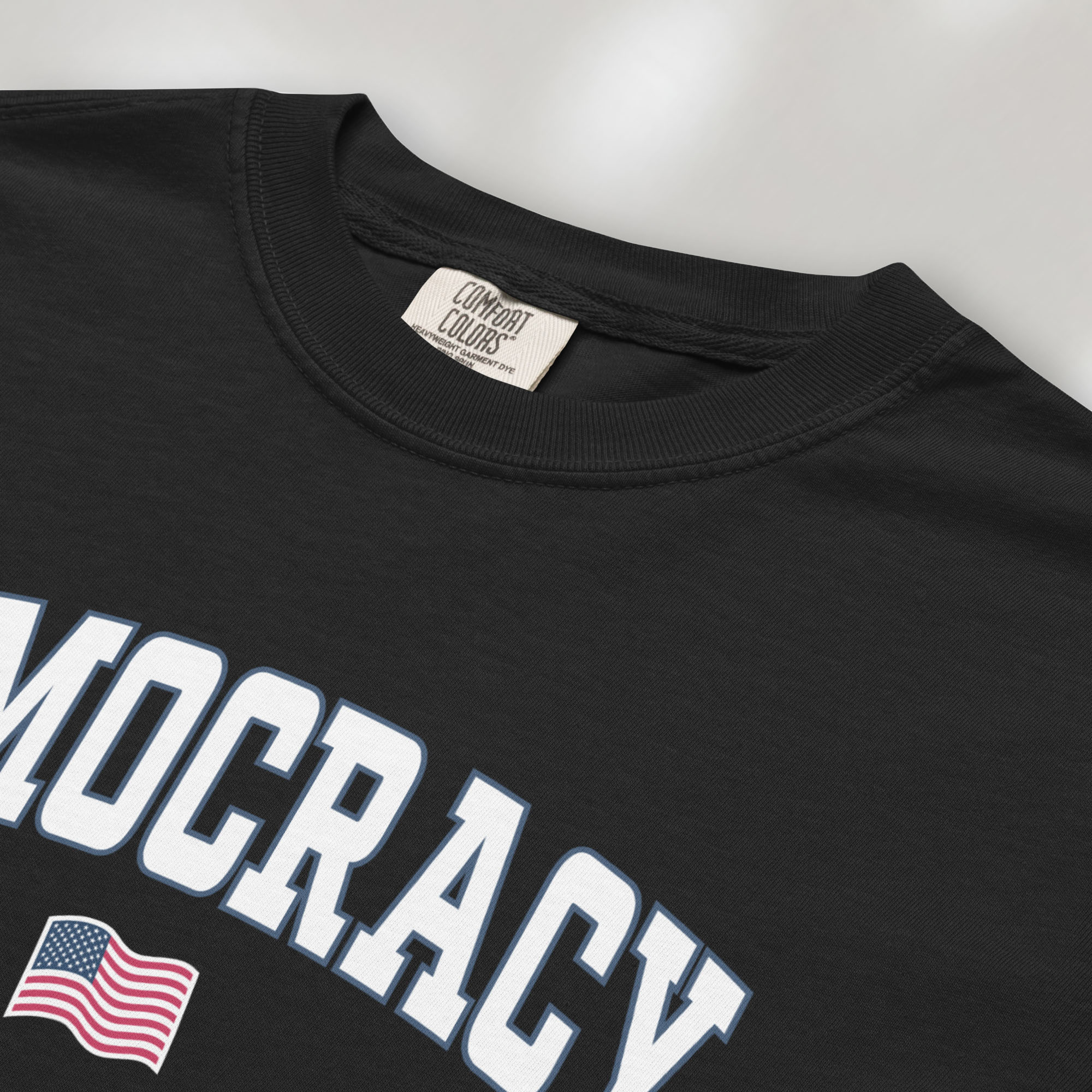 Closeup detail of black t-shirt with varsity-style graphic that says "democracy" with a USA flag beneath it.