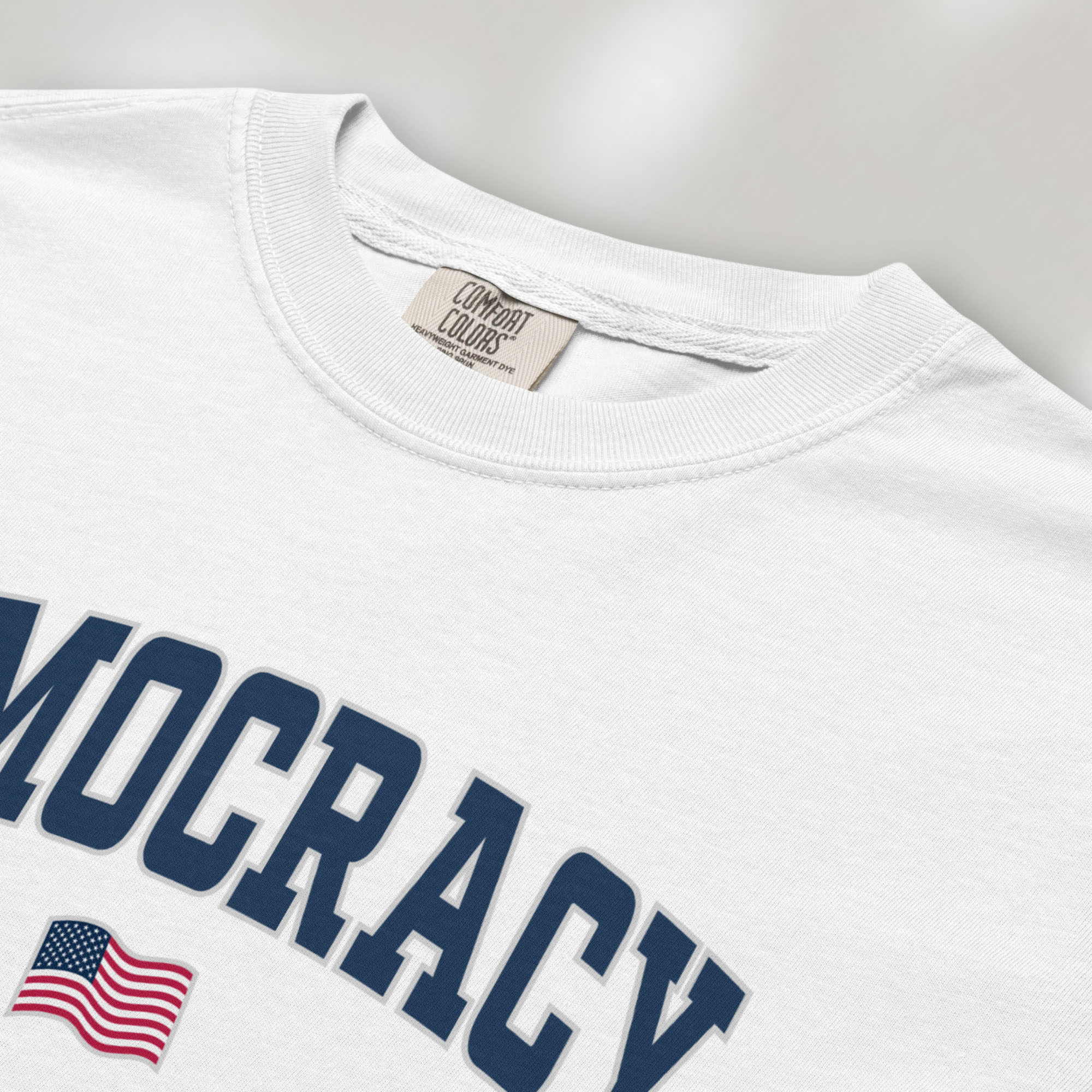 Closeup detail of white t-shirt with varsity-style graphic that says "democracy" with a USA flag beneath it.