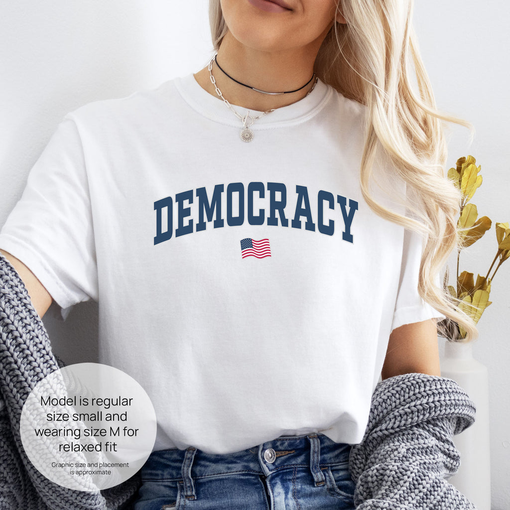 Female model wearing white t-shirt with varsity-style graphic that says "democracy" with a USA flag beneath it. Includes size and fit information for the model who is regular size small, and wearing a size M shirt for a relaxed fit.
