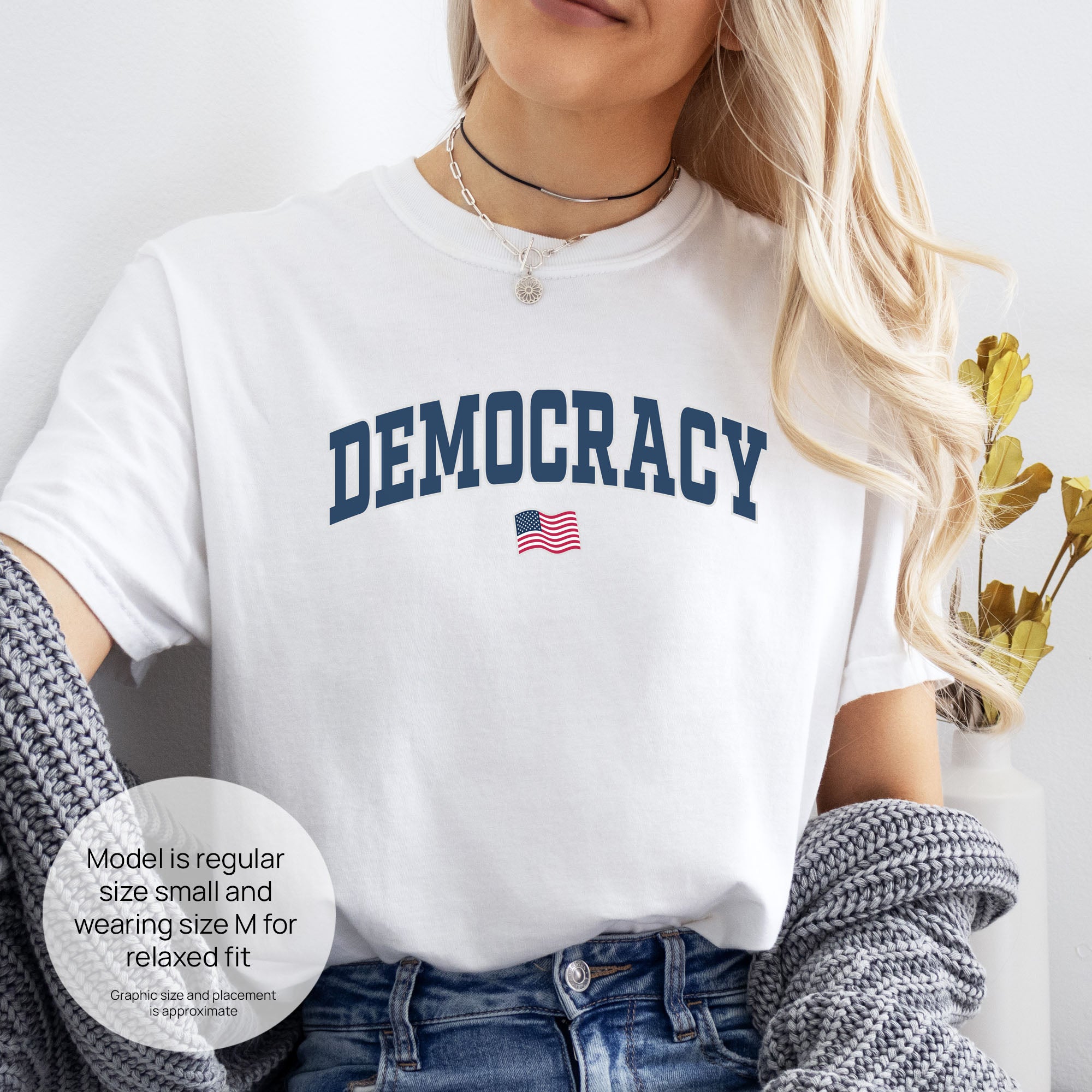 Female model wearing white t-shirt with varsity-style graphic that says "democracy" with a USA flag beneath it. Includes size and fit information for the model who is regular size small, and wearing a size M shirt for a relaxed fit.
