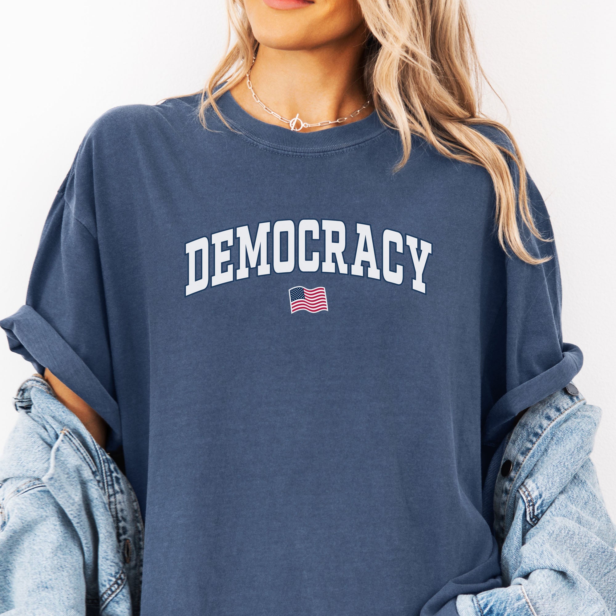 Female model wearing oversized denim t-shirt with varsity-style graphic that says "democracy" with a USA flag beneath it.
