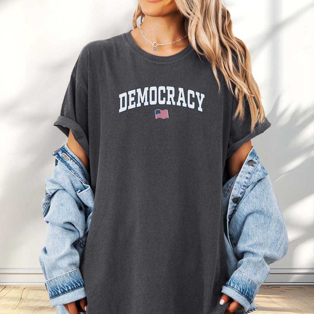 Female model wearing oversized pepper t-shirt with varsity-style graphic that says "democracy" with a USA flag beneath it.