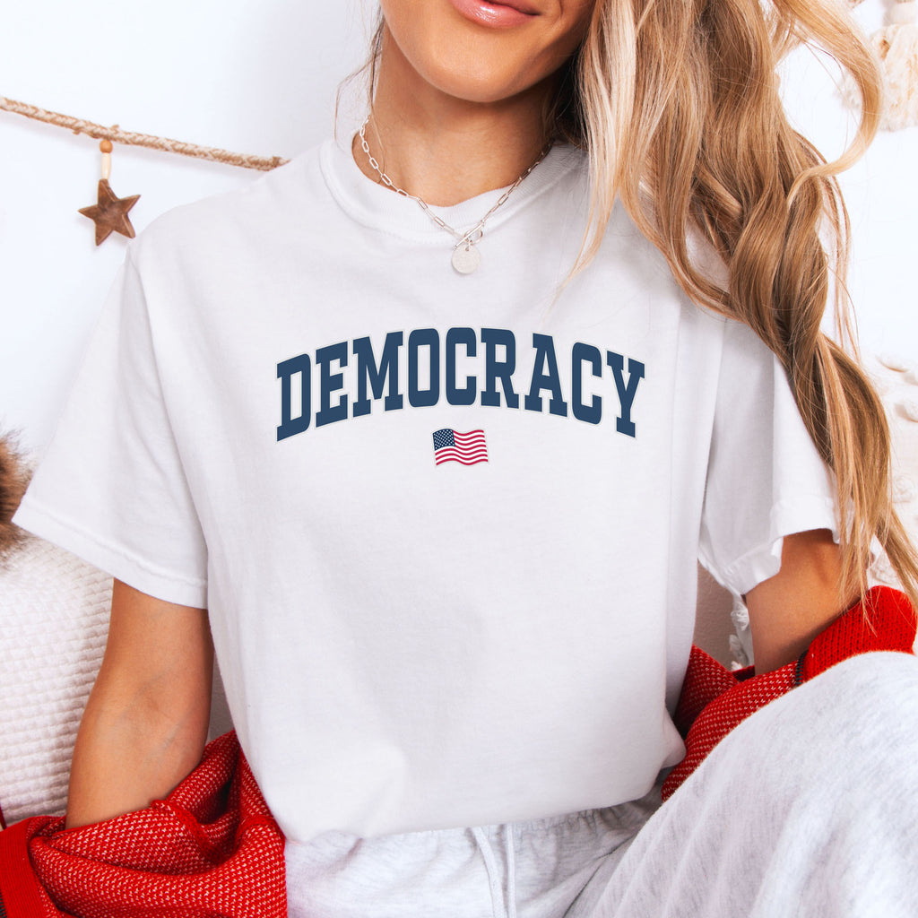 Female model wearing white t-shirt with varsity-style graphic that says "democracy" with a USA flag beneath it.
