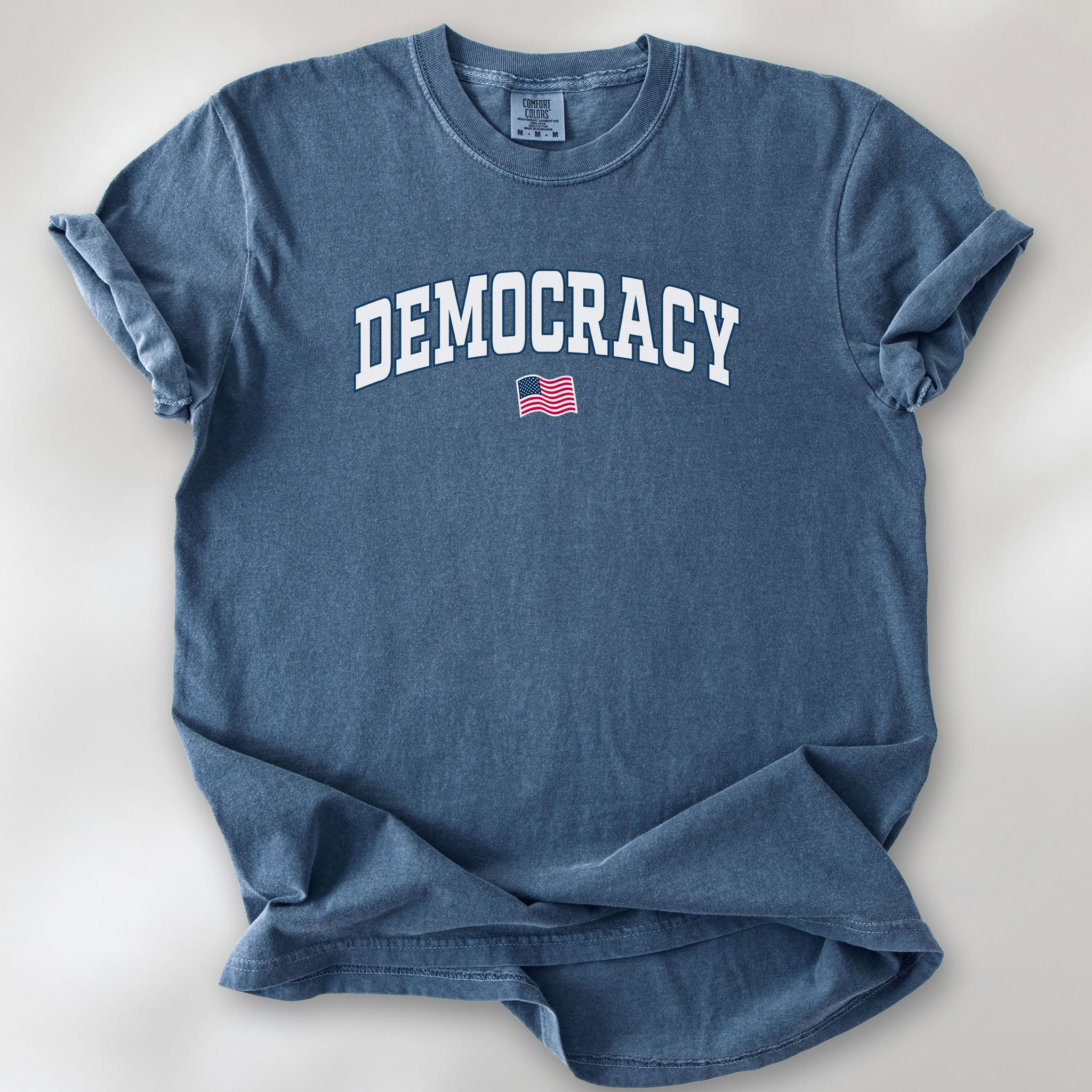 Denim t-shirt with varsity-style graphic that says "democracy" with a USA flag beneath it.