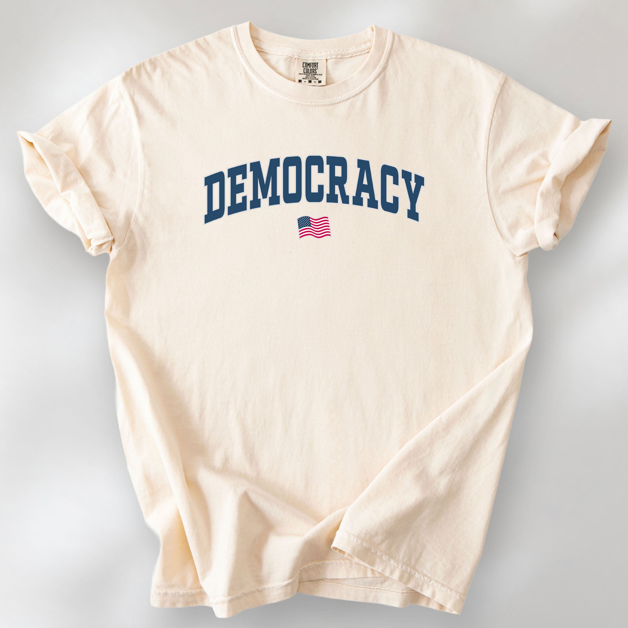 Ivory t-shirt with varsity-style graphic that says "democracy" with a USA flag beneath it.
