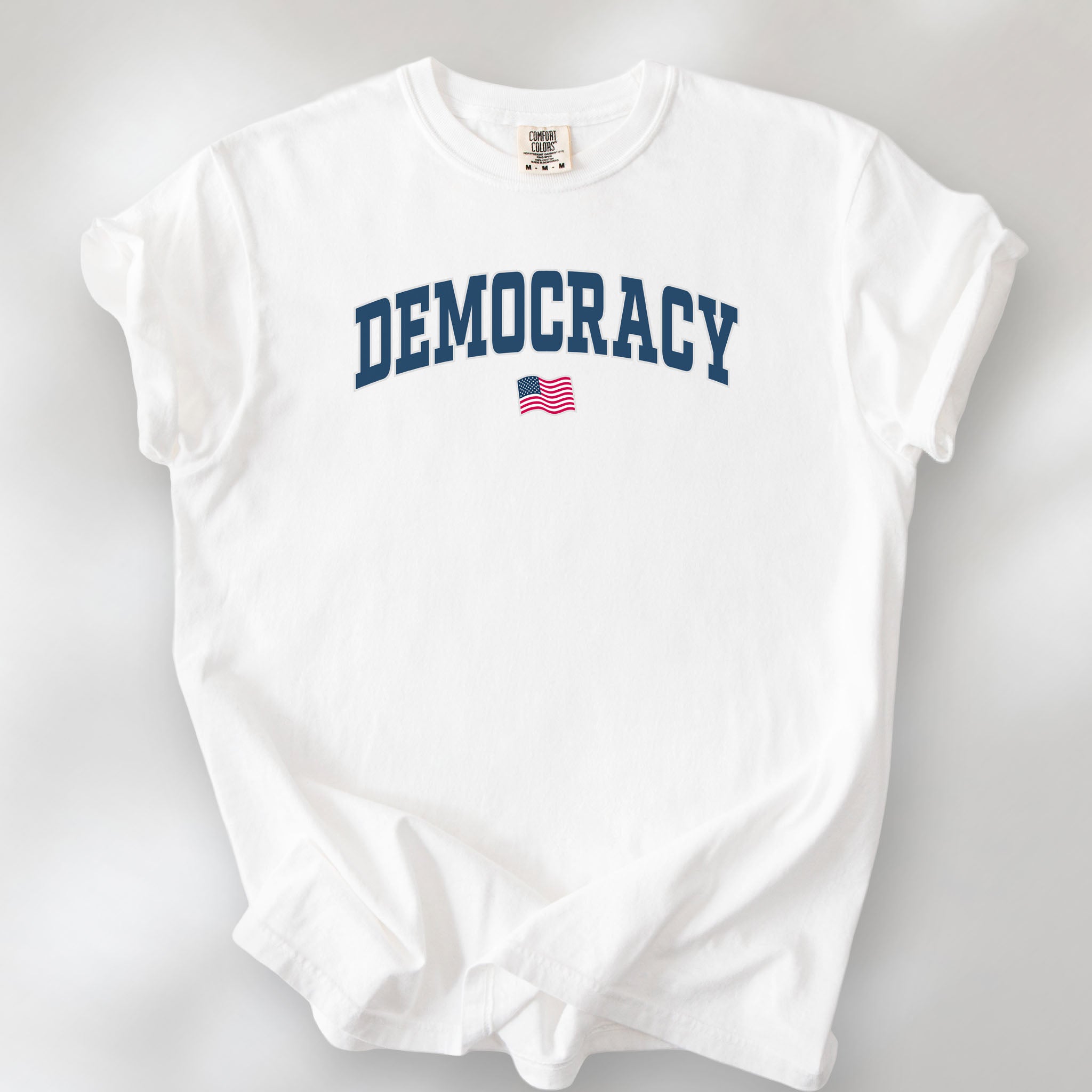 White t-shirt with varsity-style graphic that says "democracy" with a USA flag beneath it.