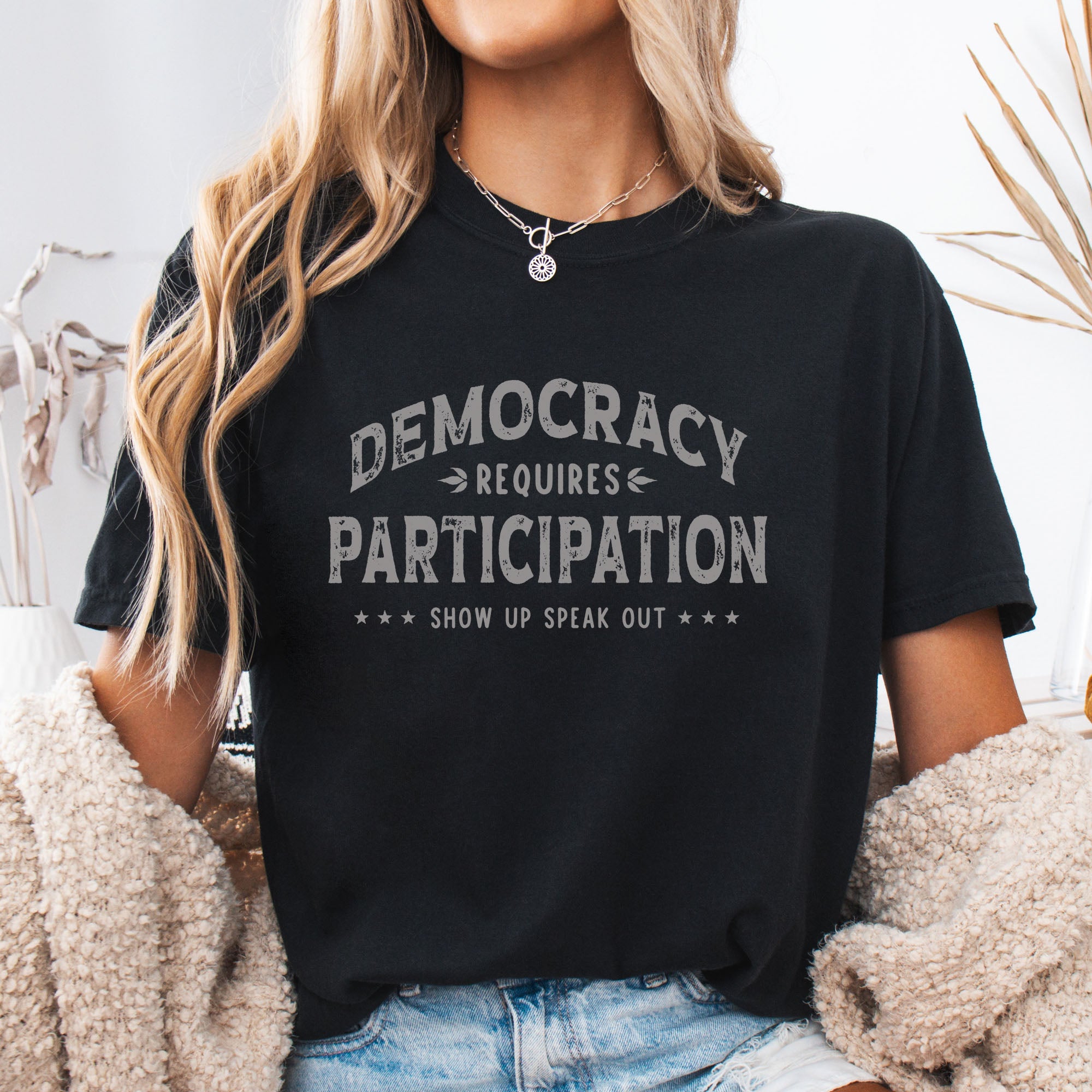 Female model wearing black t-shirt with vintage style typography reading “Democracy Requires Participation — Show Up Speak Out”.