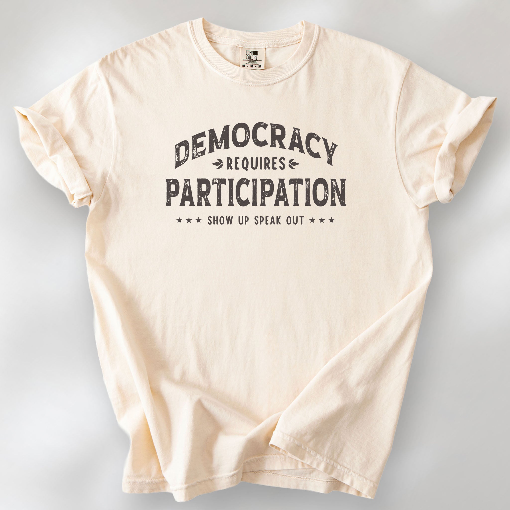Ivory t-shirt with vintage style typography reading “Democracy Requires Participation — Show Up Speak Out”.